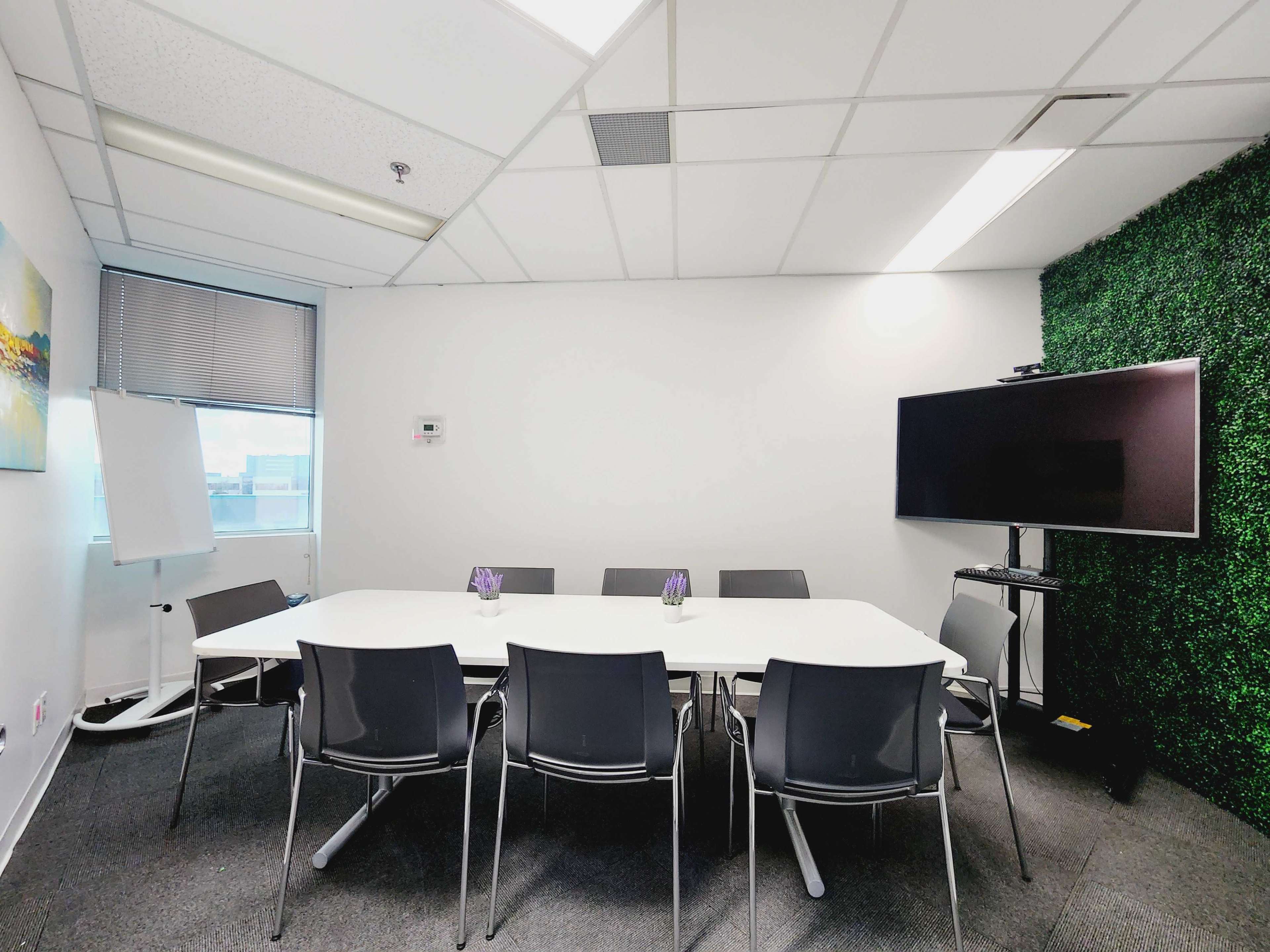The image shows a modern meeting room with a white table surrounded by gray chairs, a large television, a flip chart, and a green wall feature.