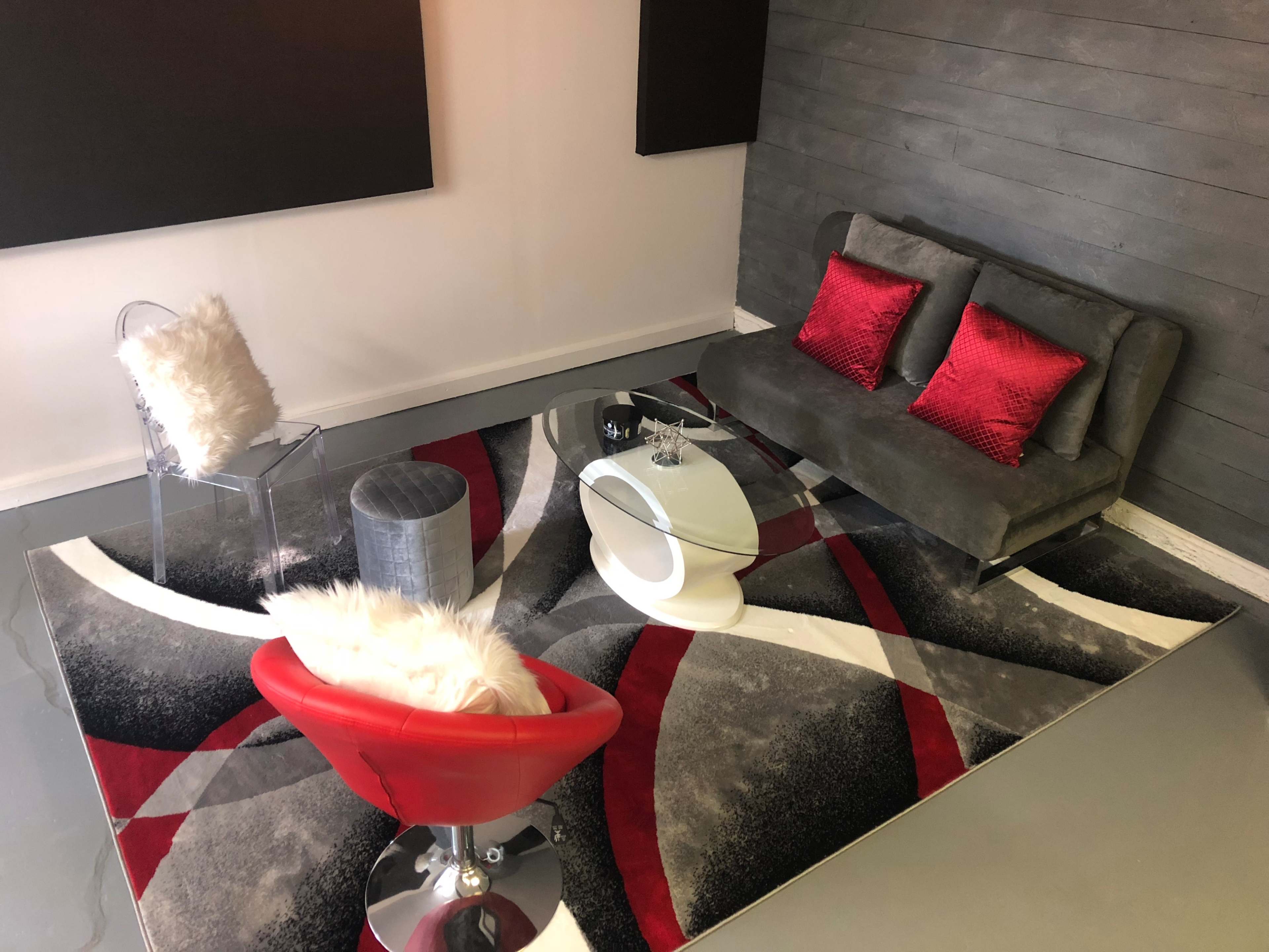 The scene features a modern lounge area with a gray sofa, red accent pillows, a glass coffee table, a silver stool, a clear chair with a white pillow, and a patterned area rug.