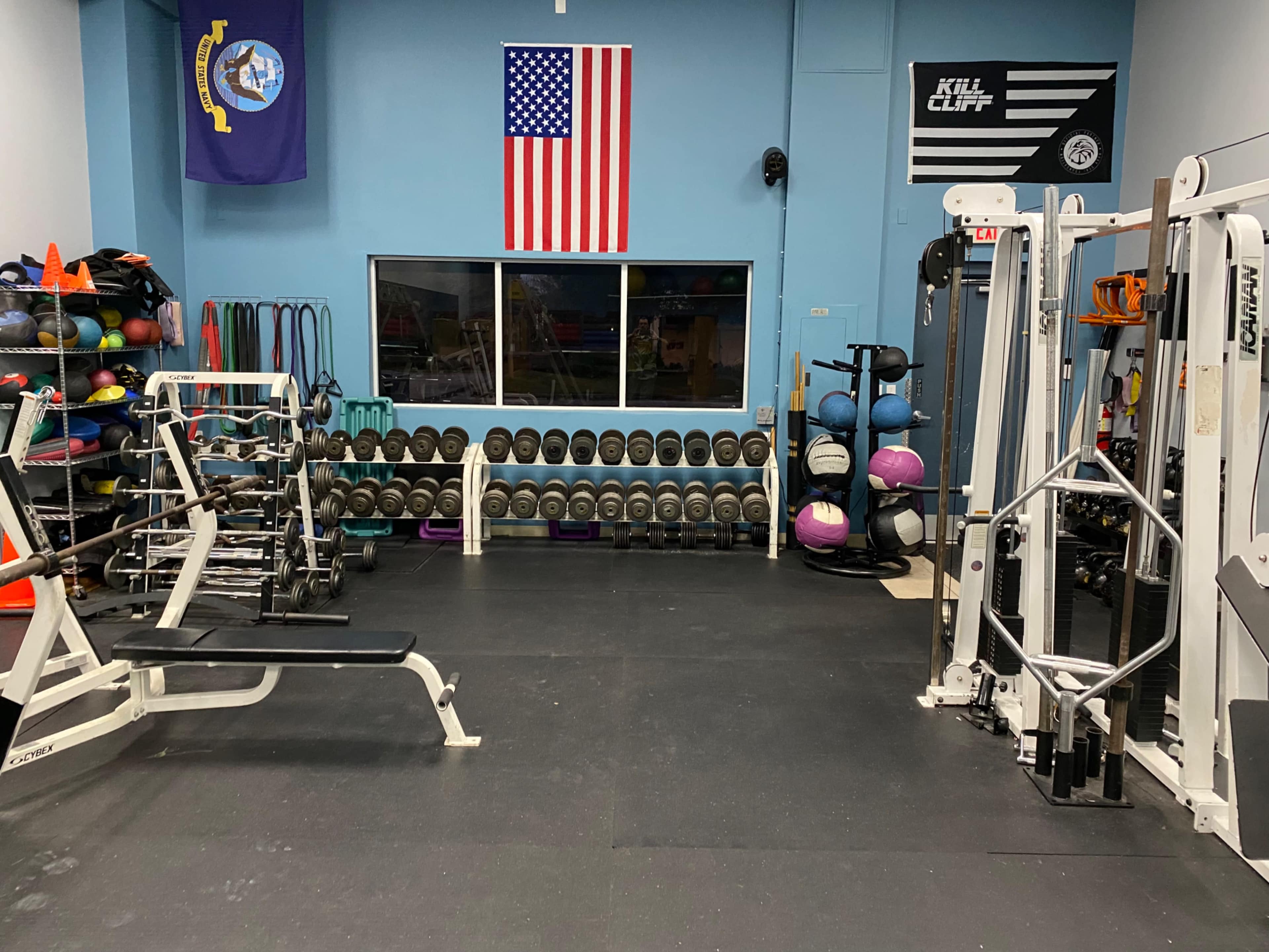 A gym interior with weightlifting equipment, exercise balls, and free weights lined up against the wall, adorned with American and military flags.
