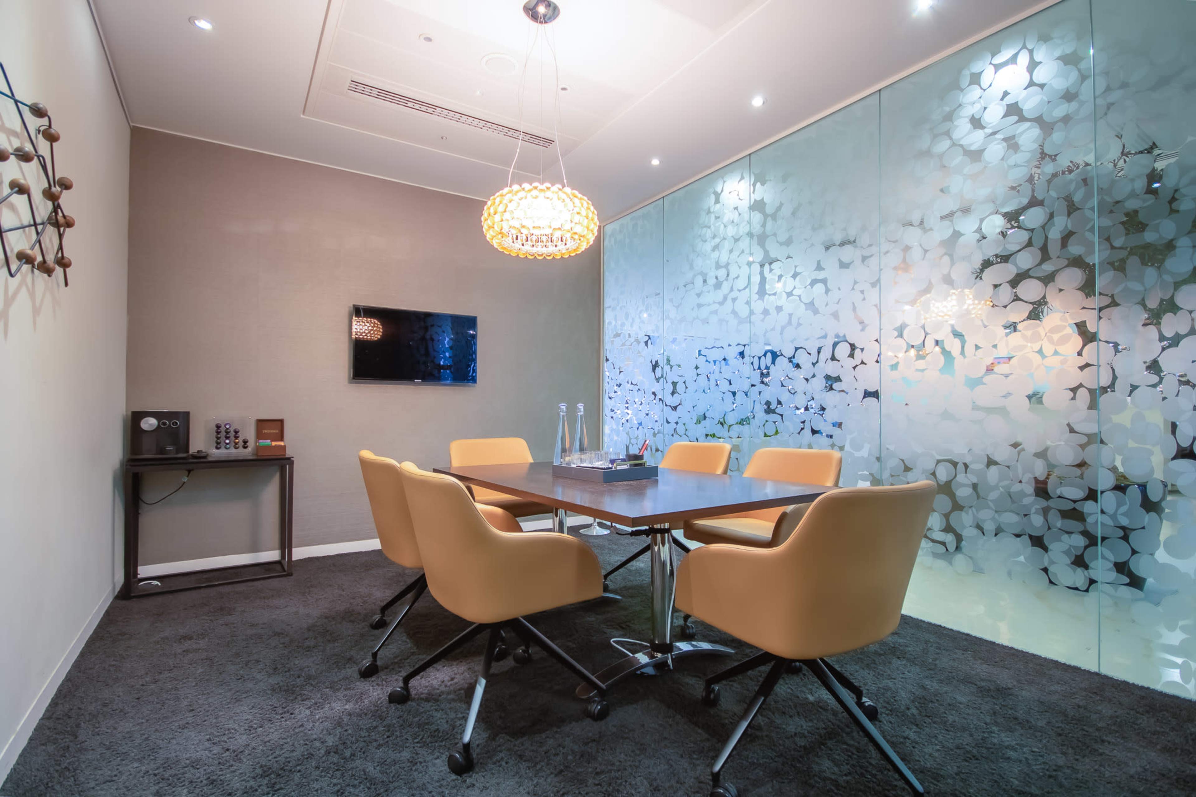 A modern meeting room features a large table surrounded by six orange chairs, with frosted glass walls and a wall-mounted television.