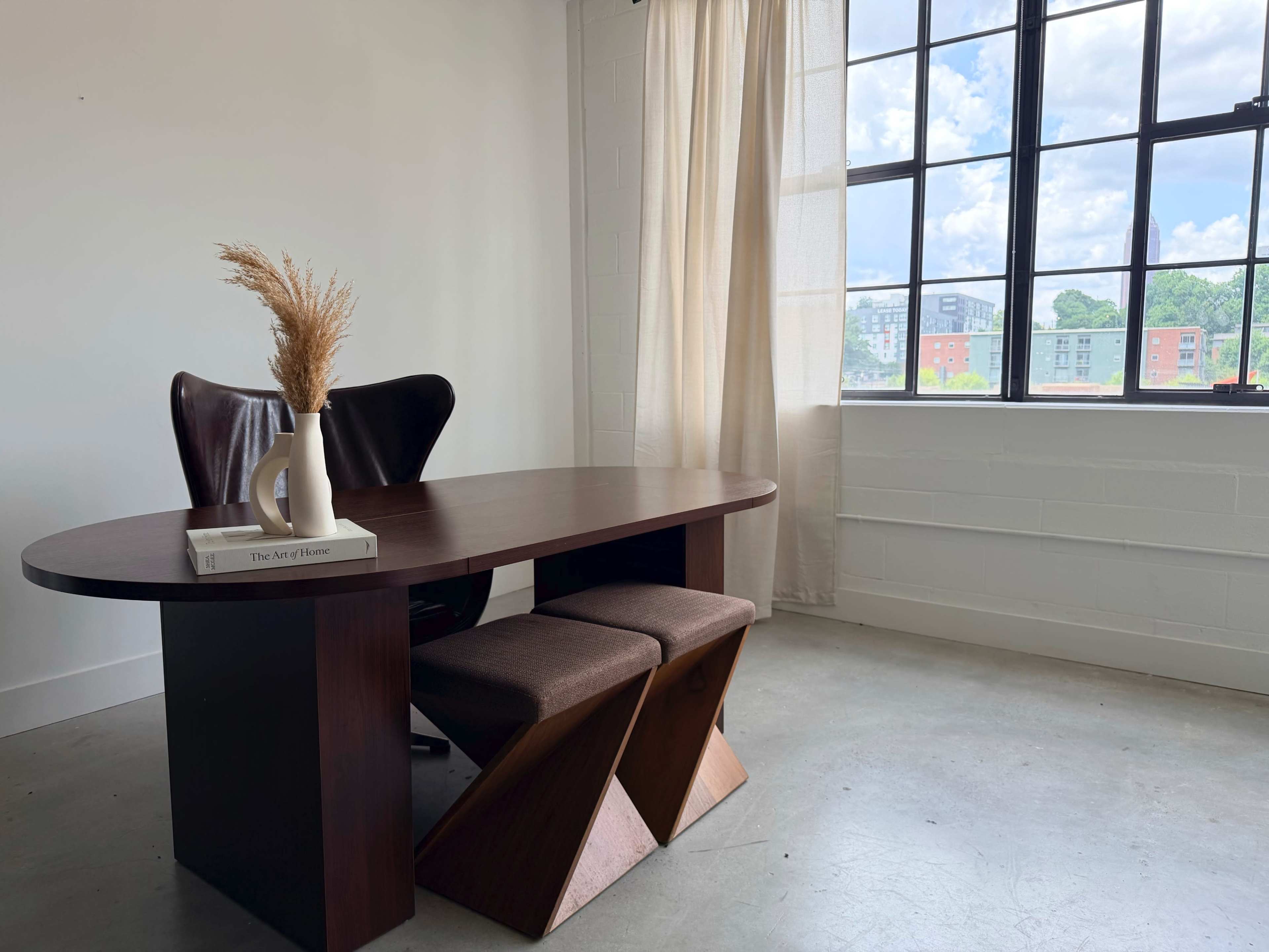 A modern office space features a curved wooden desk with a vase of dried plants, two cushioned stools, and large windows allowing natural light to fill the room.