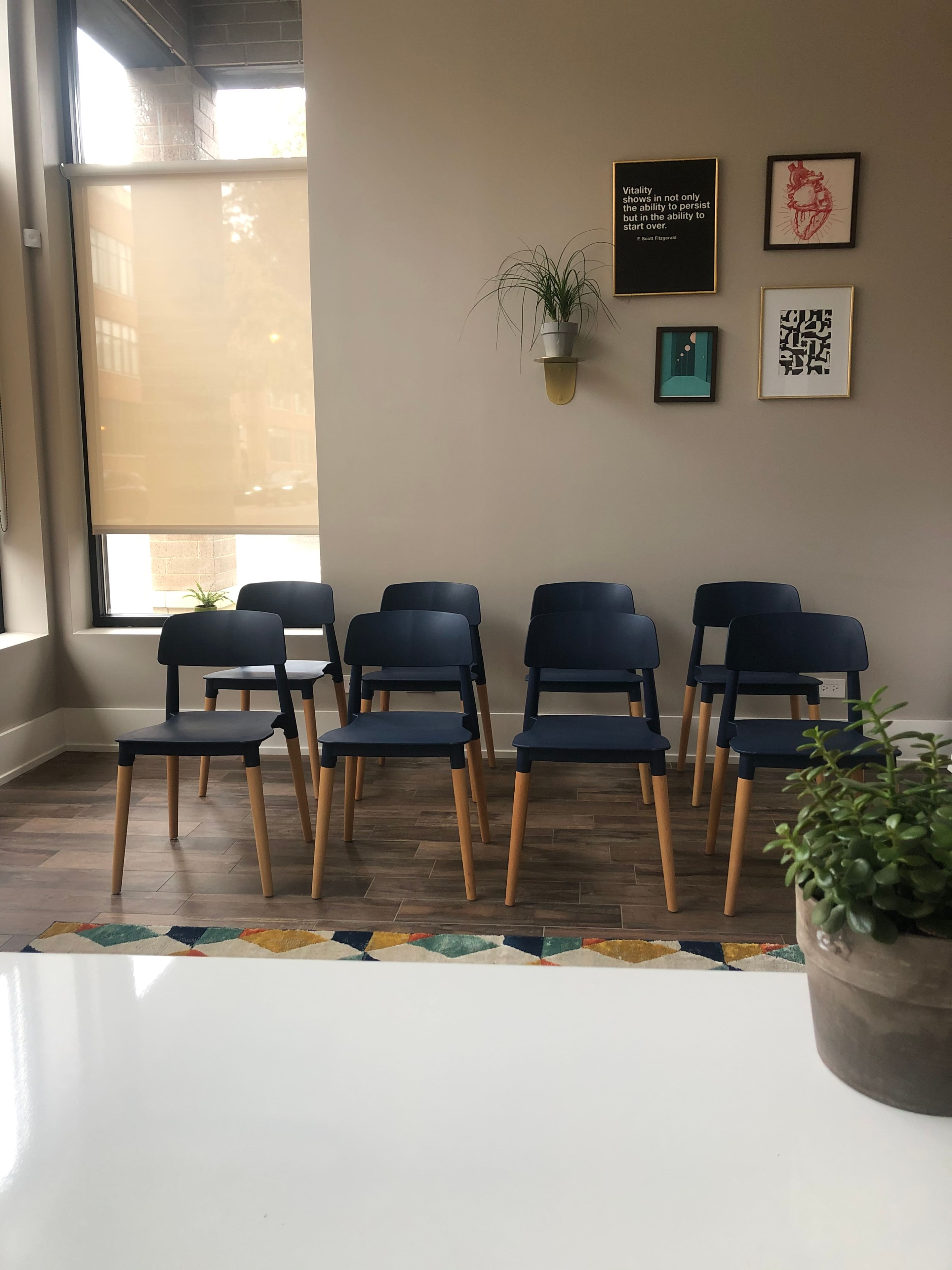 A row of blue chairs with wooden legs is positioned against a wall, accompanied by framed art and a small potted plant on a table in the foreground.