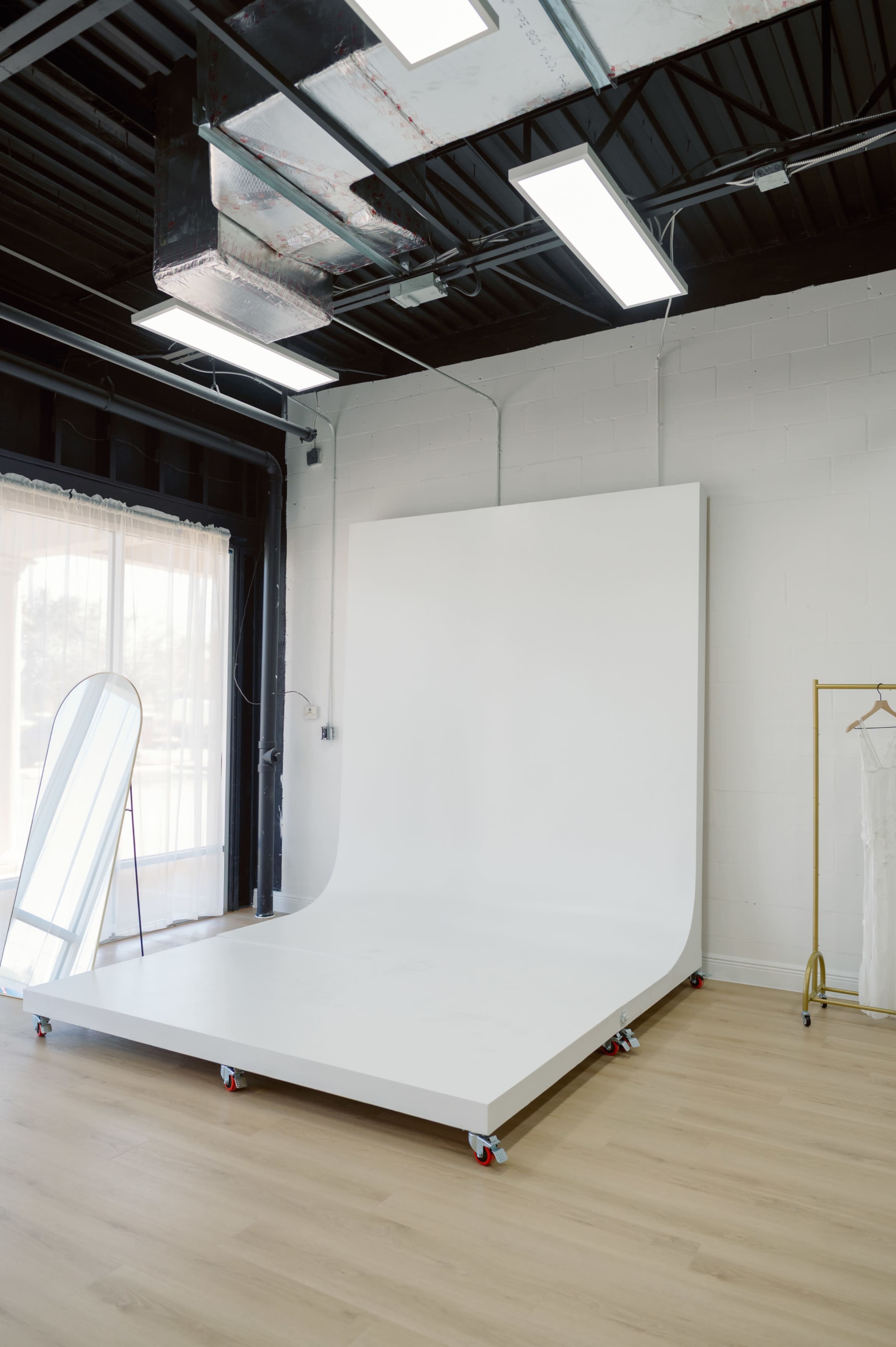 The image features a minimalist photography studio with a large white backdrop on wheels, a full-length mirror, and a clothing rack, all set against a simple wooden floor.