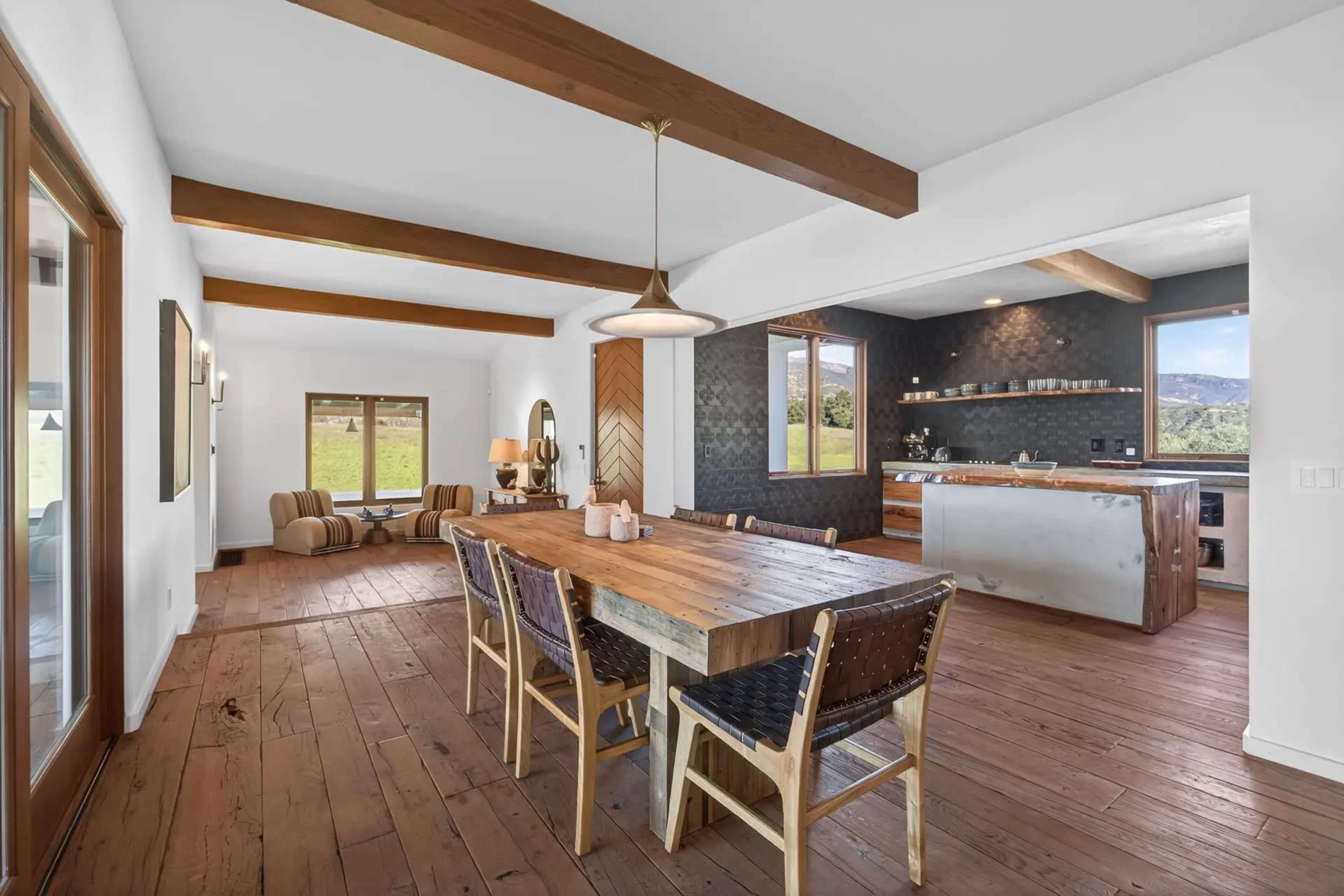 The image shows a spacious dining area with a wooden table, surrounded by chairs, and an open kitchen in the background featuring modern appliances and a large island.