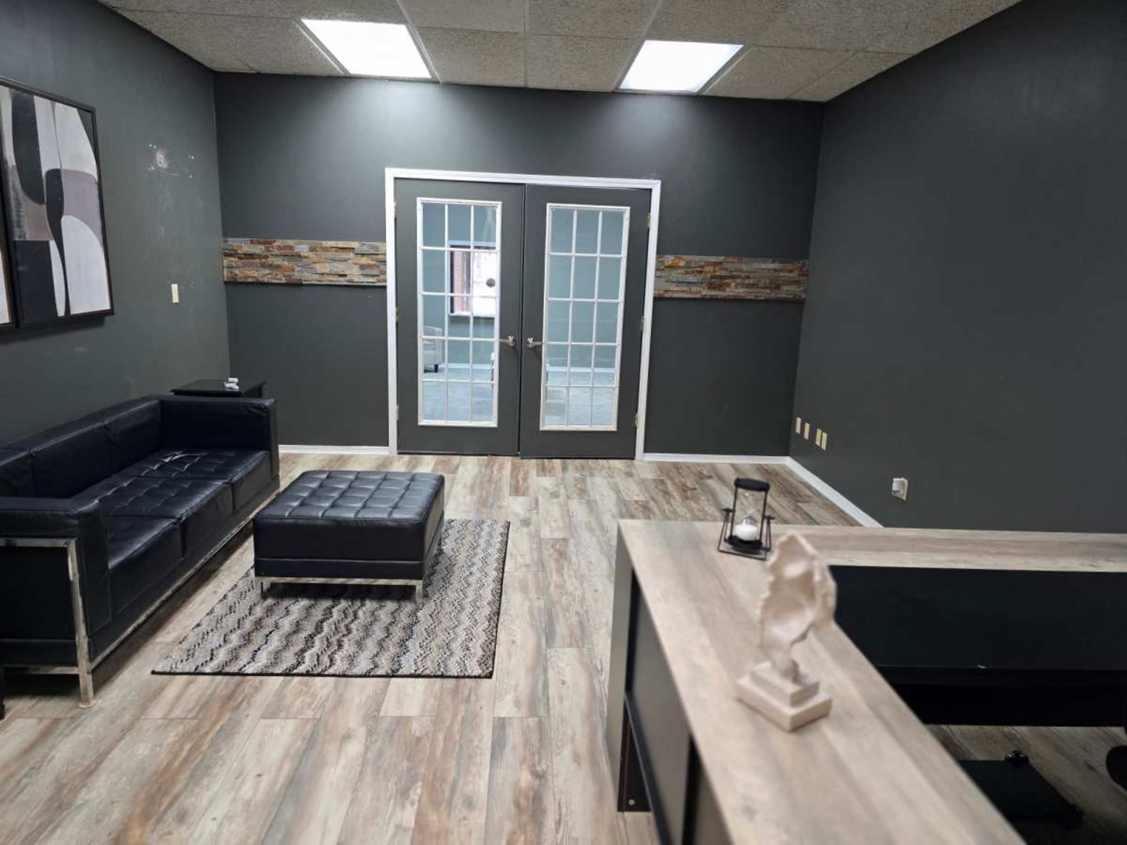 The image shows a modern waiting area with a black sectional sofa, a square coffee table, and double doors leading to another room.