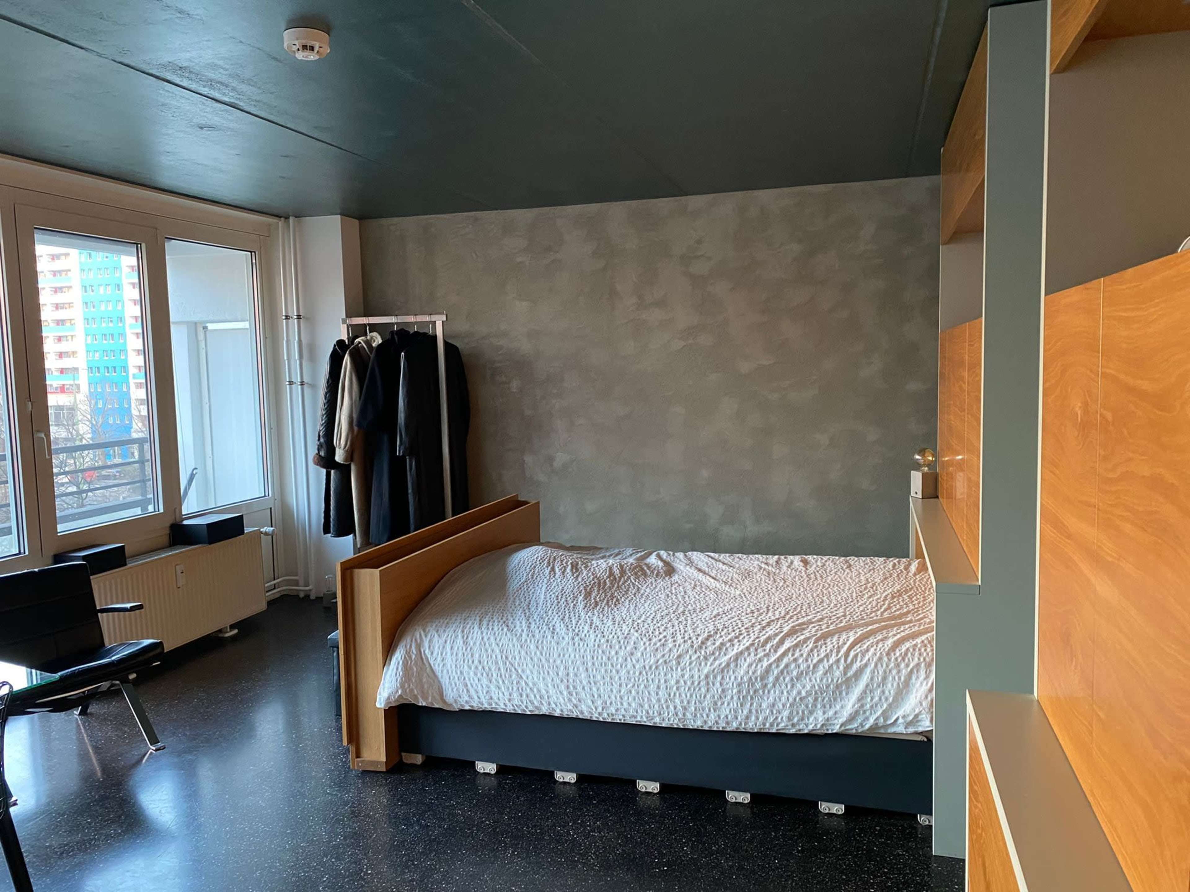 The image shows a simple bedroom with a bed, a chair, and a clothing rack against a textured wall, with large windows letting in natural light.