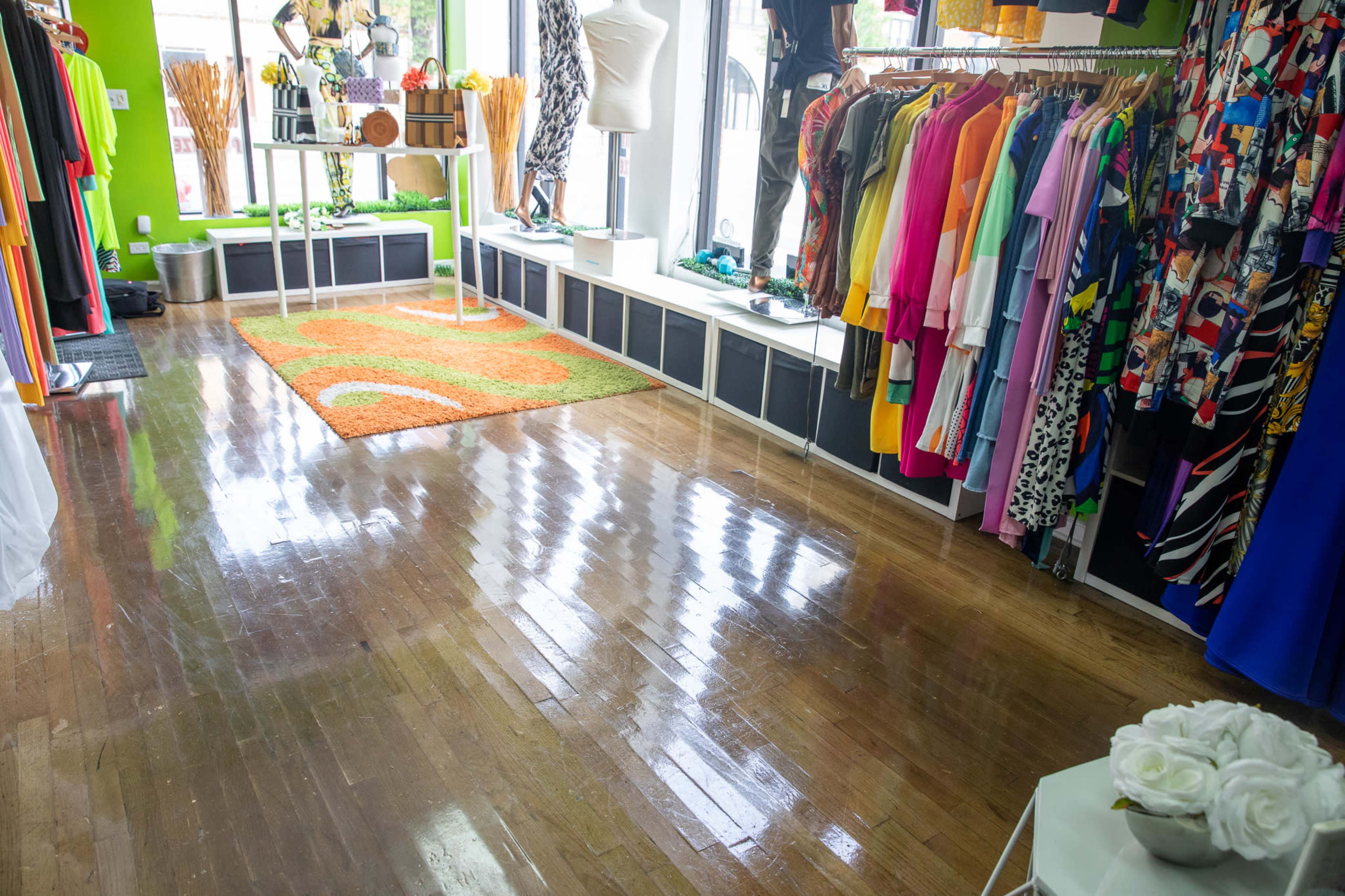 The image shows a brightly colored boutique with clothing displays, a decorative rug, and polished wooden floors reflecting natural light.