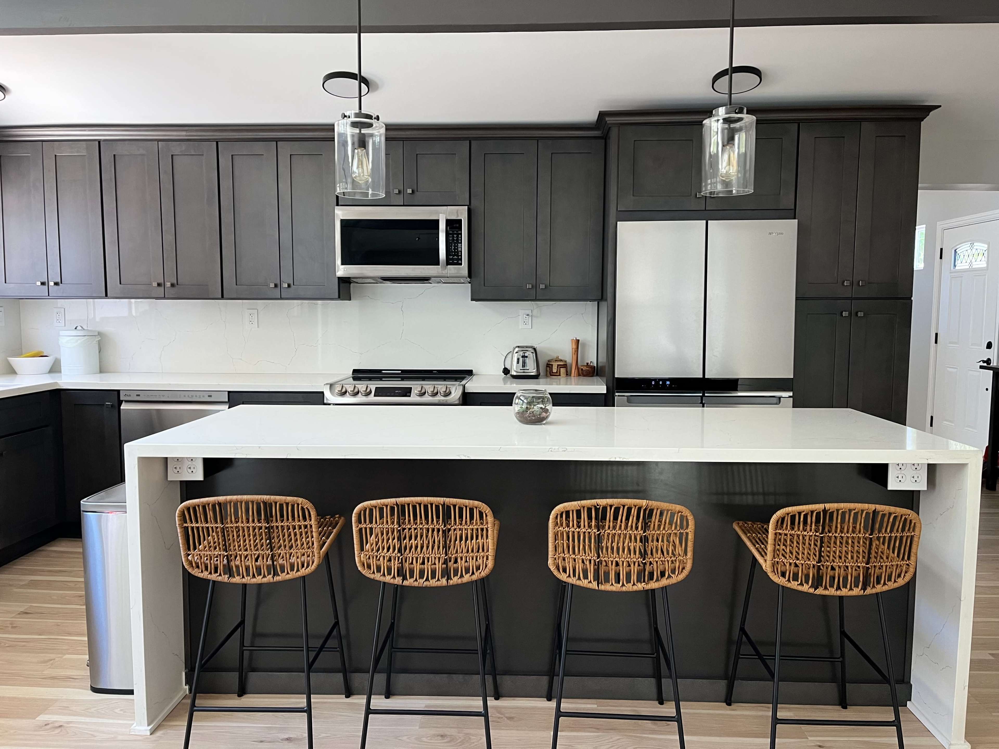 The kitchen features dark cabinets, stainless steel appliances, and a white countertop with four woven bar stools.