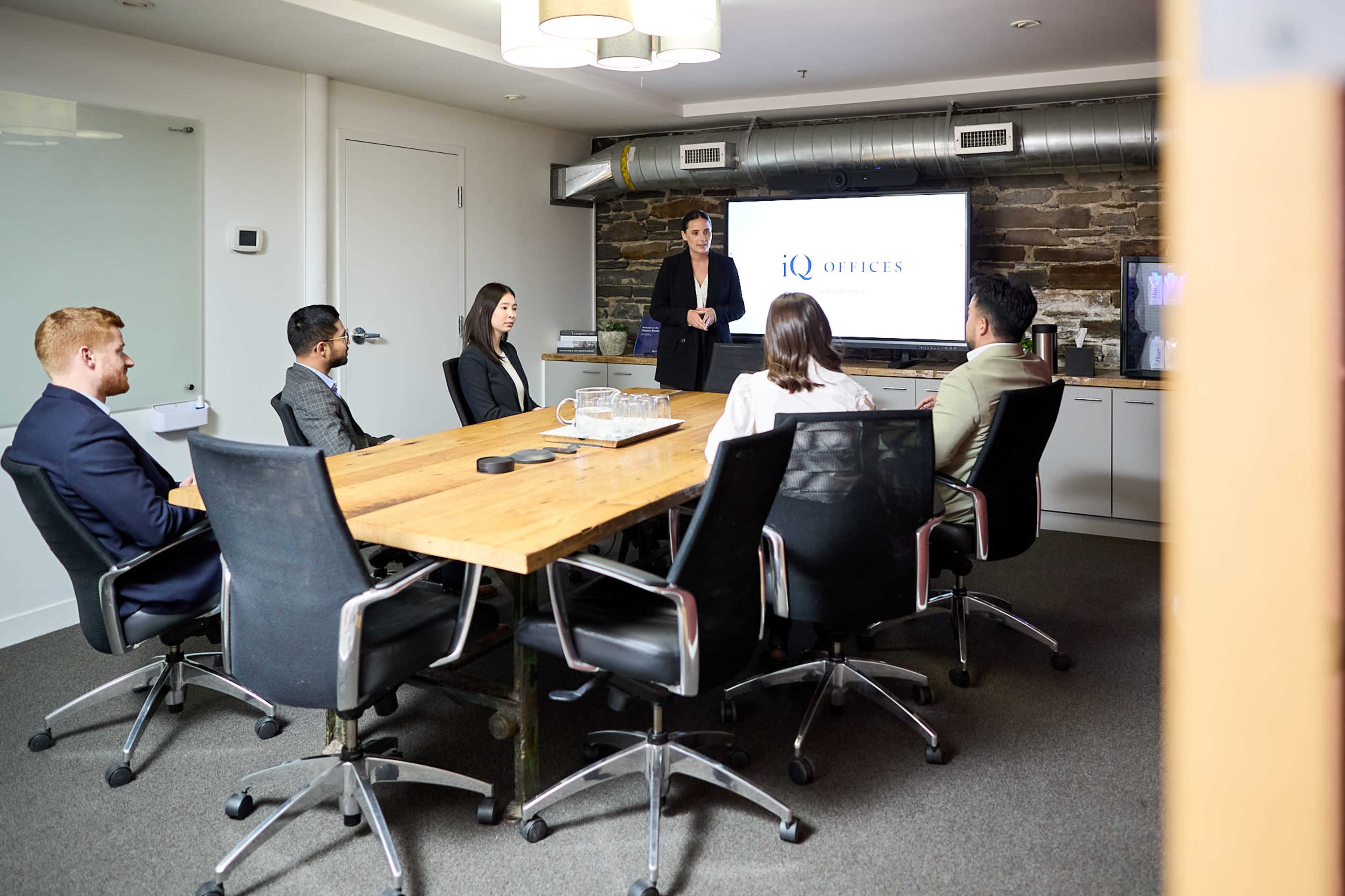 A group of five people in business attire is seated around a wooden conference table, while a sixth person stands at the front of the room presenting to them.