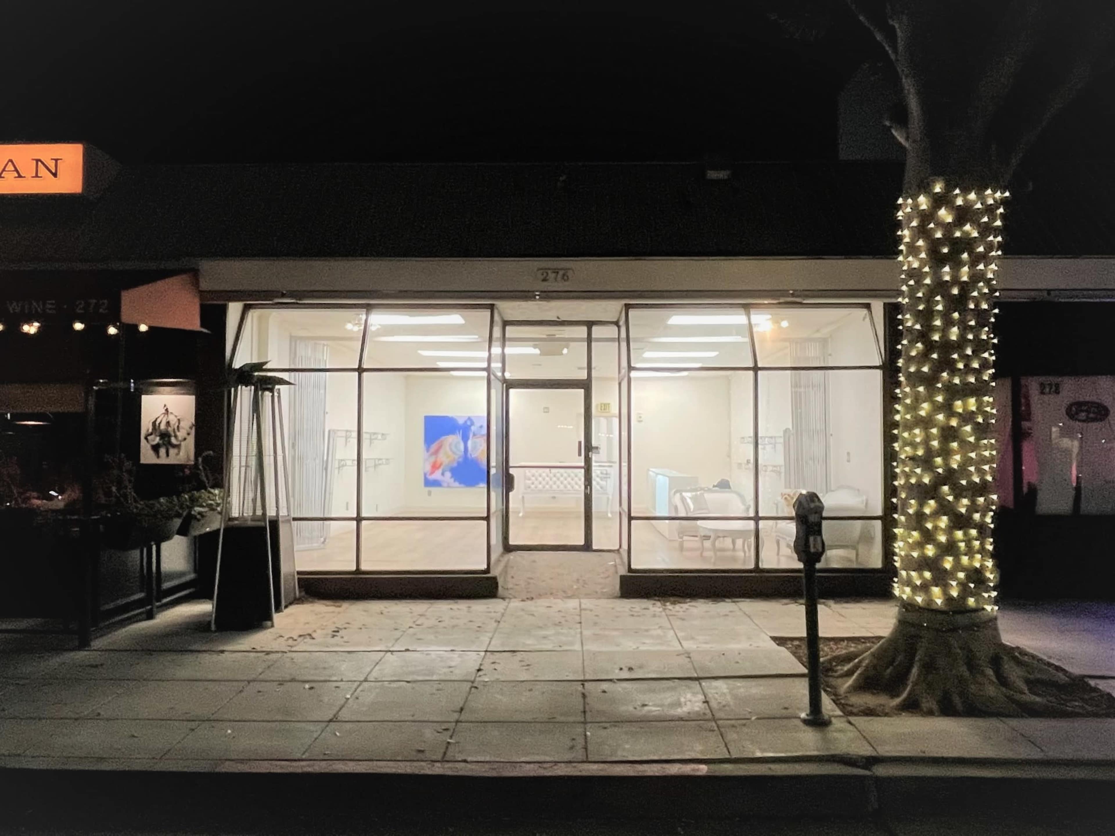 A brightly lit storefront with glass windows showcases a minimalistic interior, while a tree wrapped in string lights stands nearby.