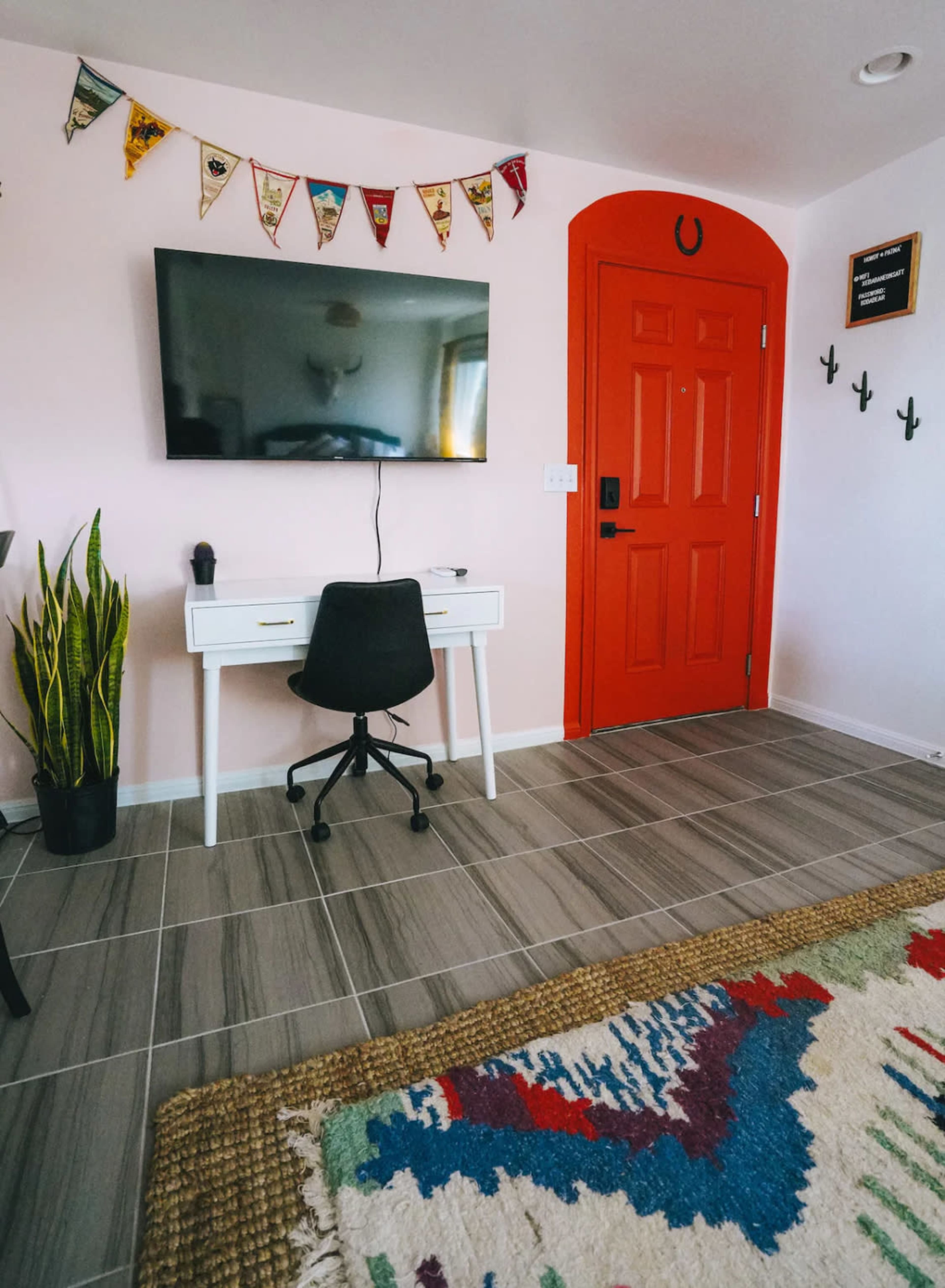 A modern workspace features a white desk and chair in front of a large wall-mounted TV, with a vibrant red door and decorative pennants overhead.