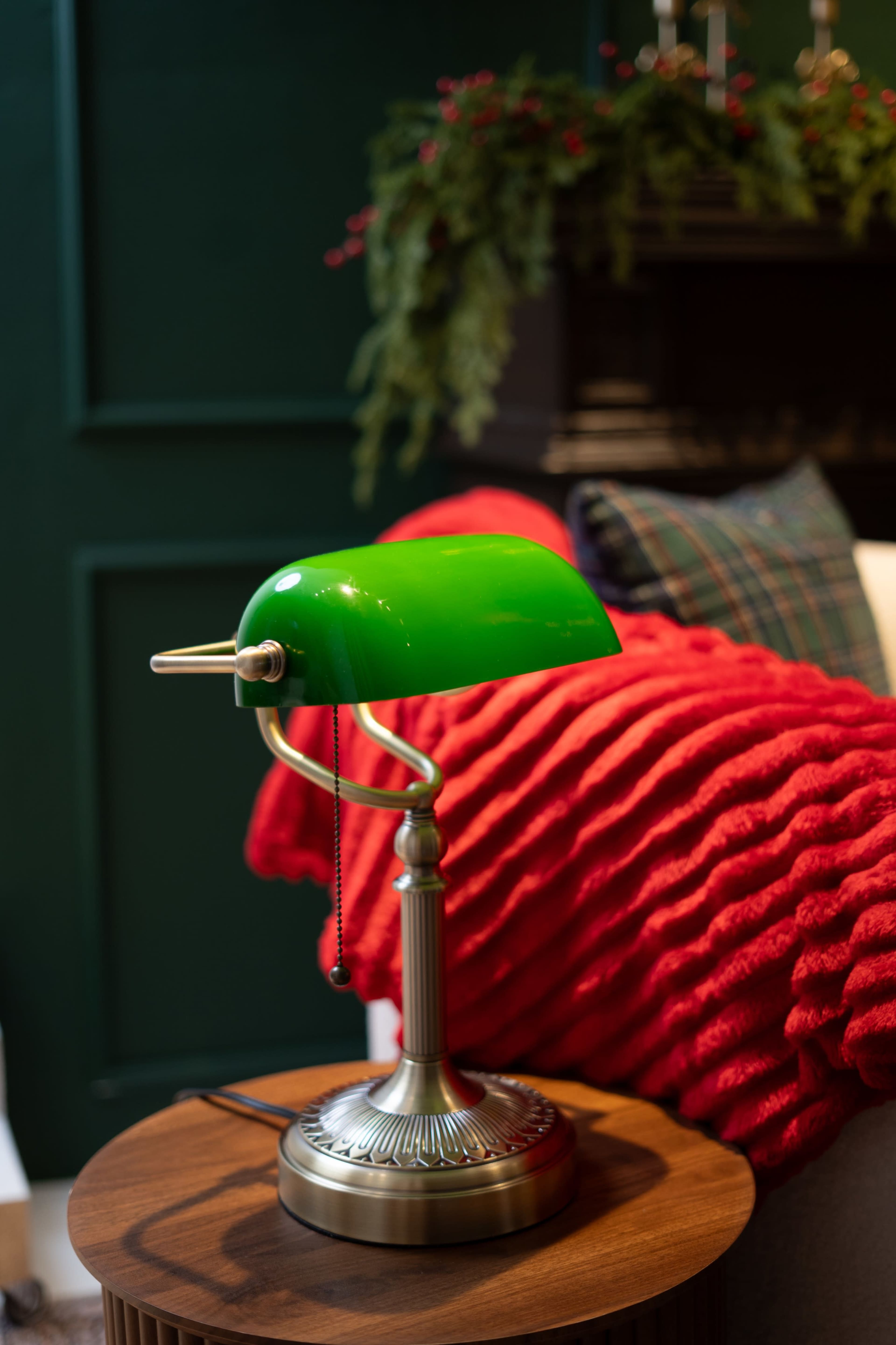 A green desk lamp with a brass base sits on a wooden table next to a red blanket, in front of a dark green wall and a cozy seating area.