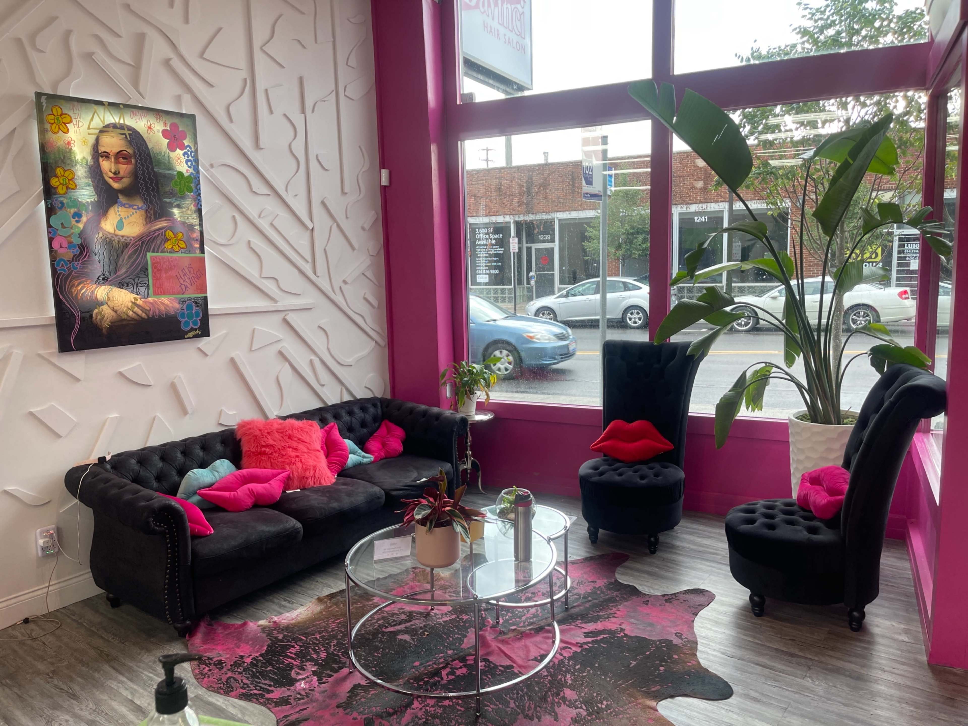 A colorful room featuring a black couch adorned with pink cushions, a round glass table, and a large plant, positioned near a bright window displaying a street view.
