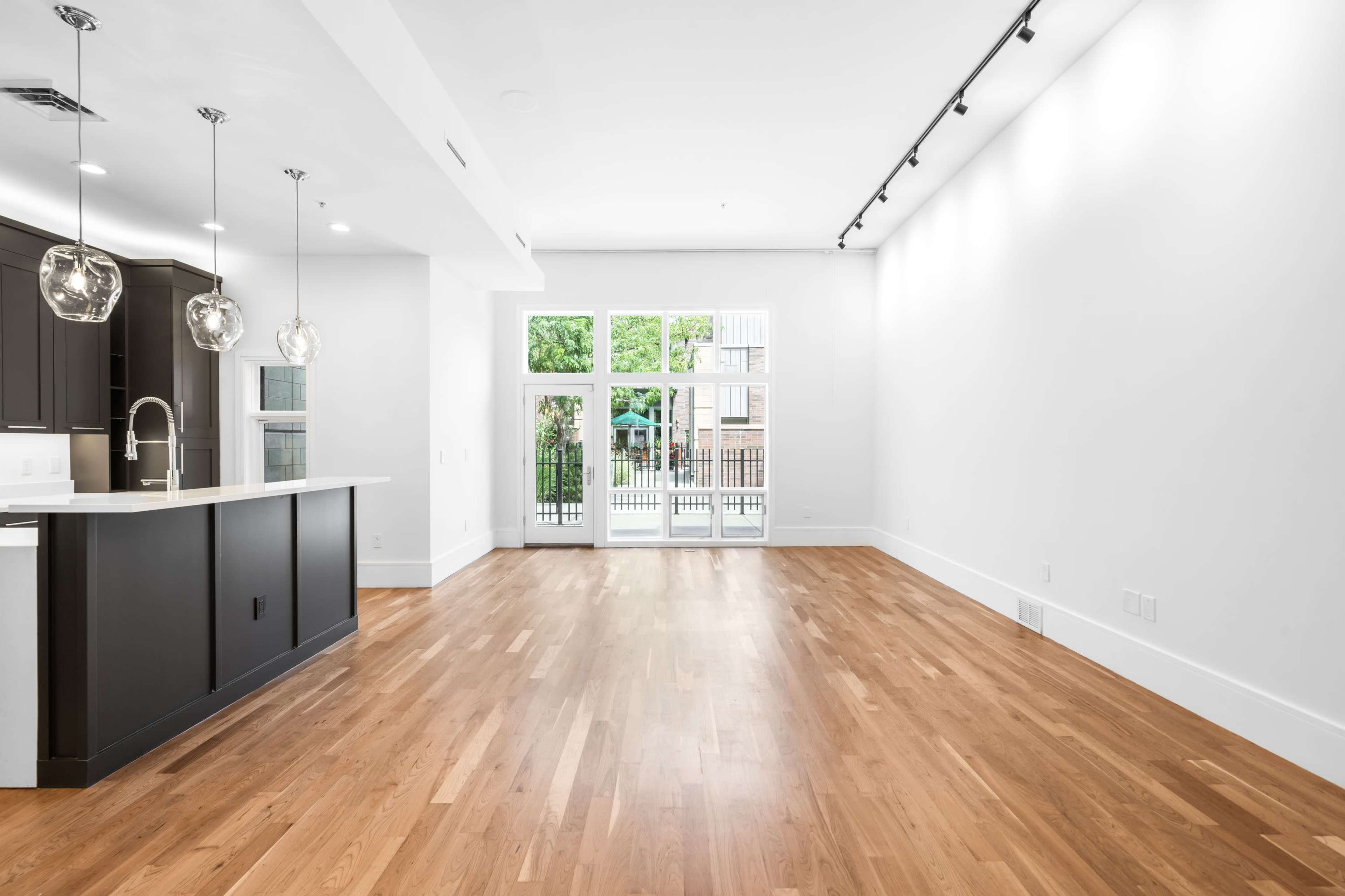 The image shows a spacious, modern interior with hardwood floors, large windows, and a kitchen island.