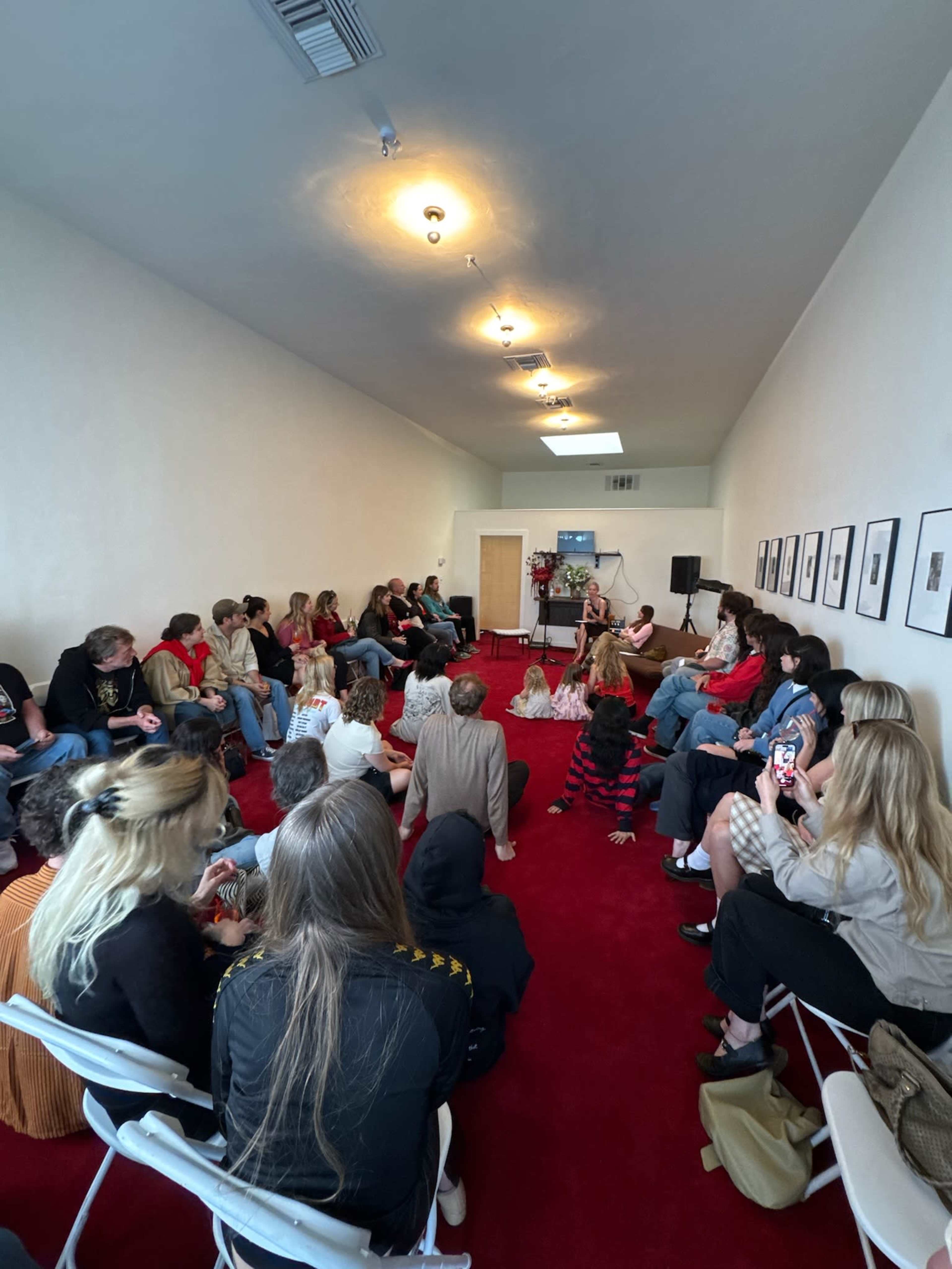 A group of people is seated on a red carpeted floor in a narrow room, facing a speaker at the front, with photographs hanging on the walls.
