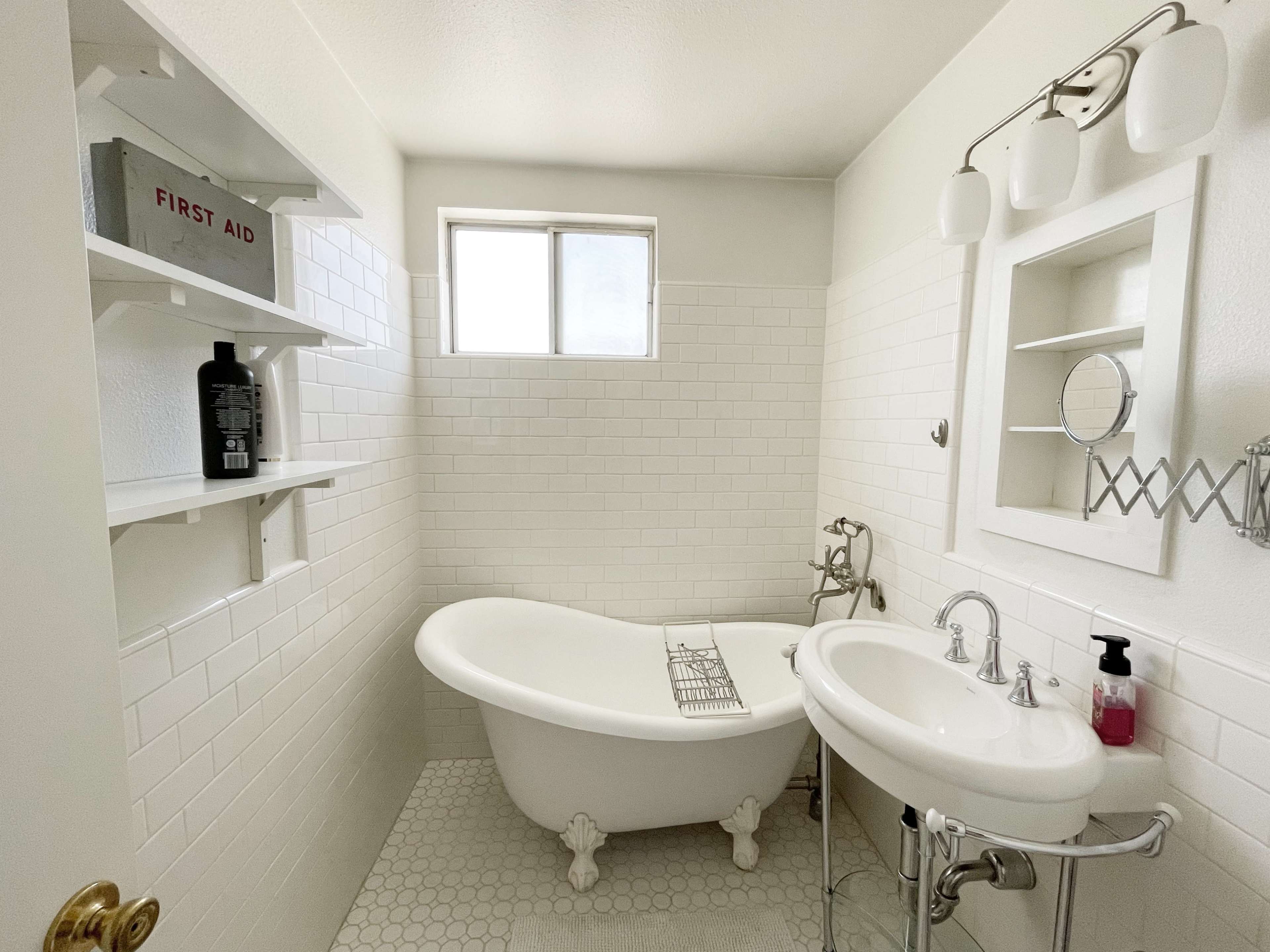 The image shows a bathroom featuring a freestanding bathtub, a sink, and tiled walls with a window allowing natural light.