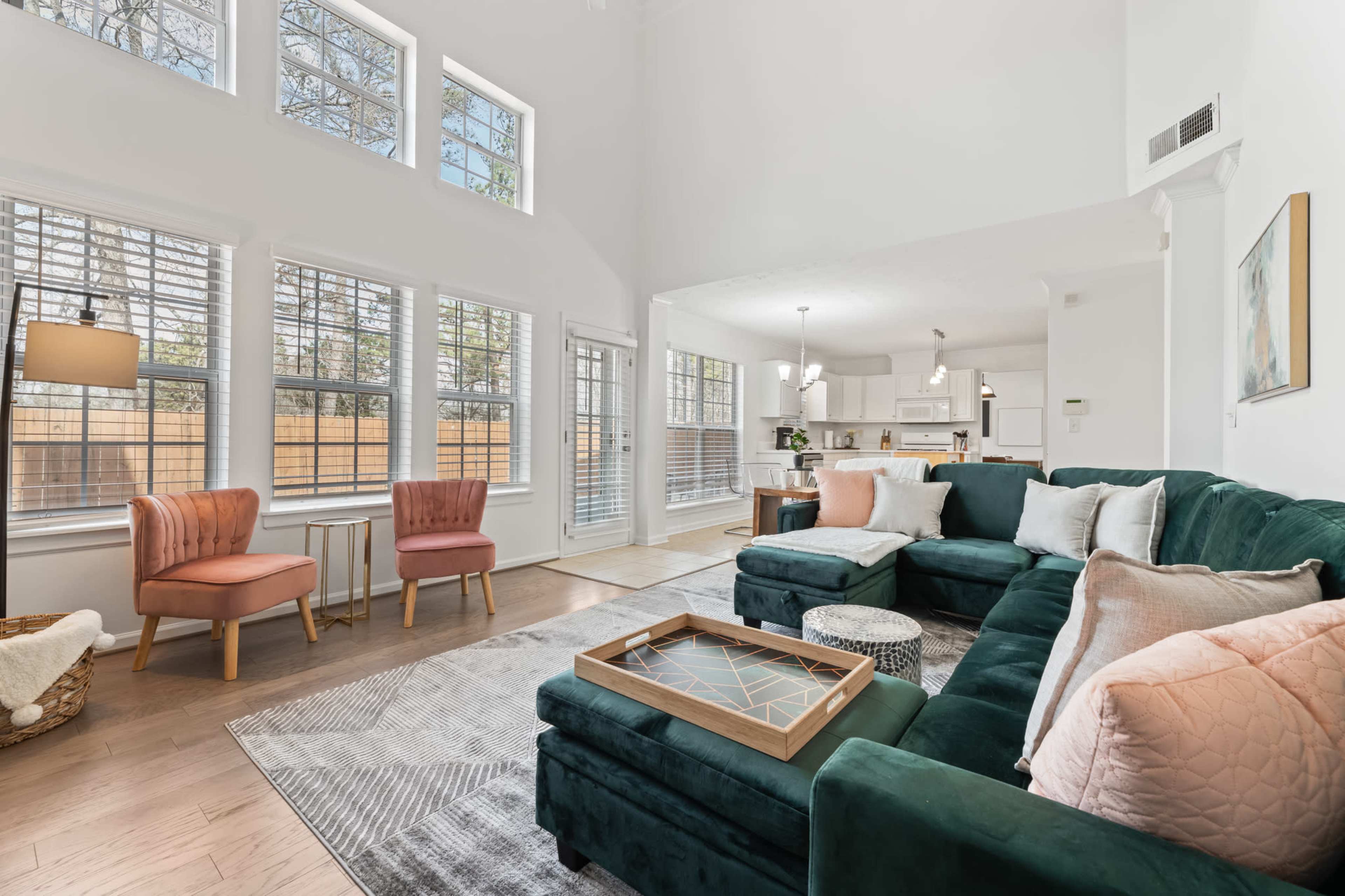 The living room features a green sectional sofa, two blush pink chairs, and large windows allowing natural light to fill the space.