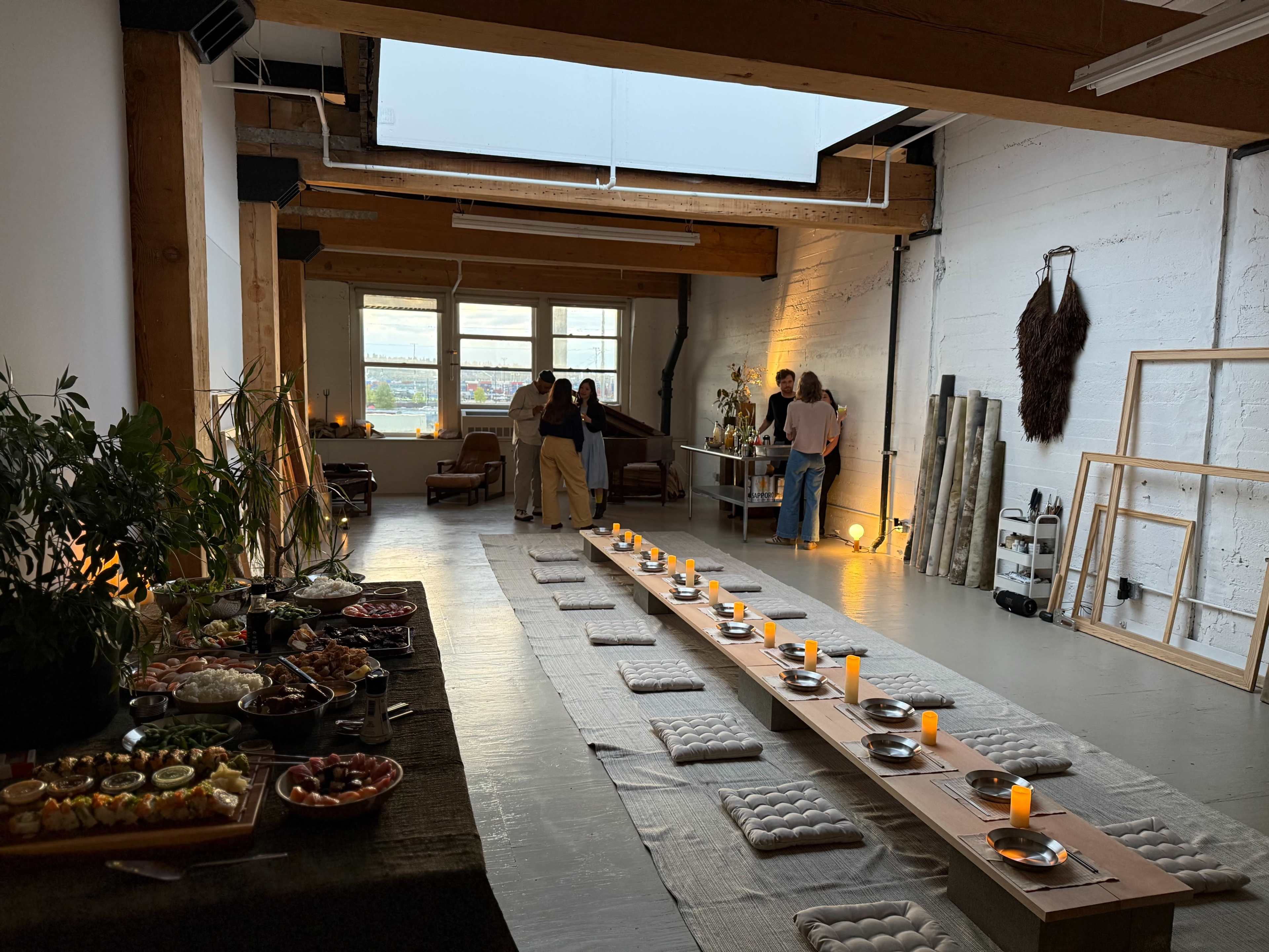 A spacious, well-lit room features a long dining setup with cushions and candles, surrounded by a variety of food laid out on a table.