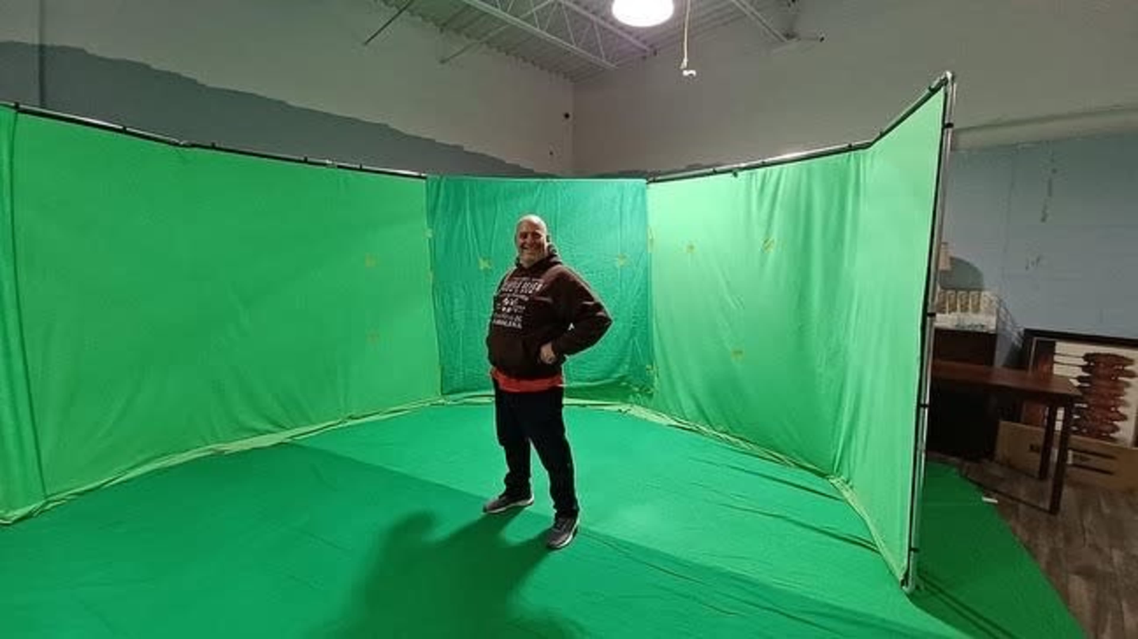 A man stands inside a green screen setup in a room with minimal furnishings.