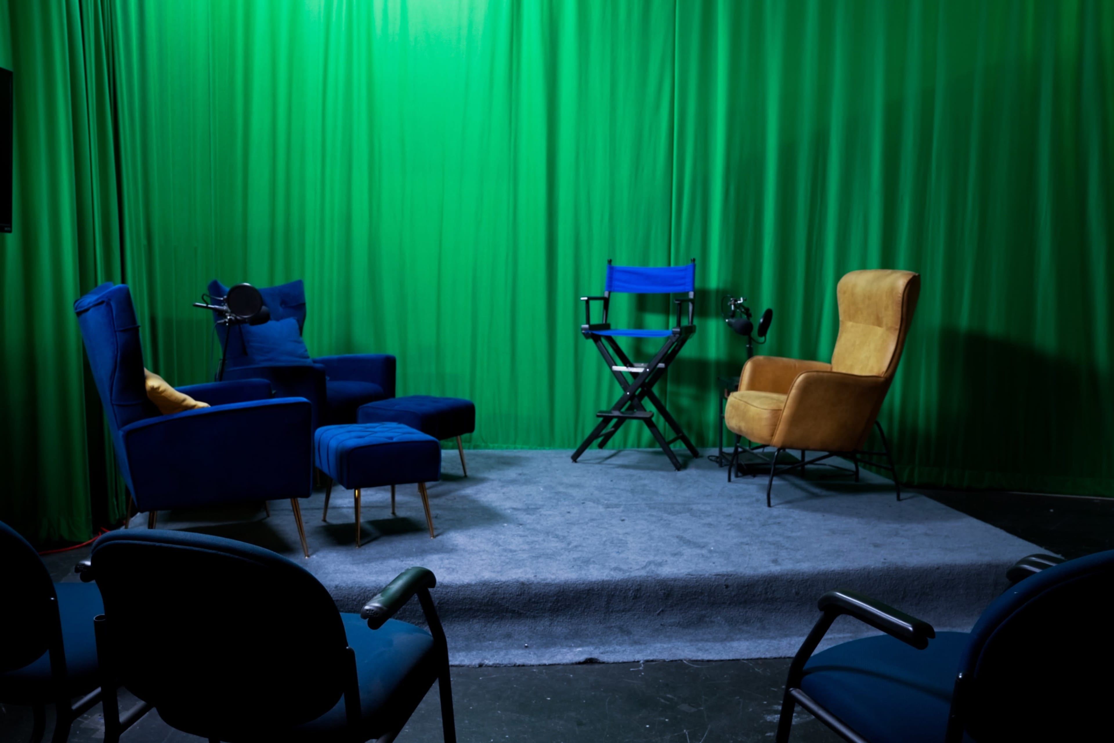 The image shows a studio setup with blue chairs, a yellow armchair, a director's chair, and a green backdrop.