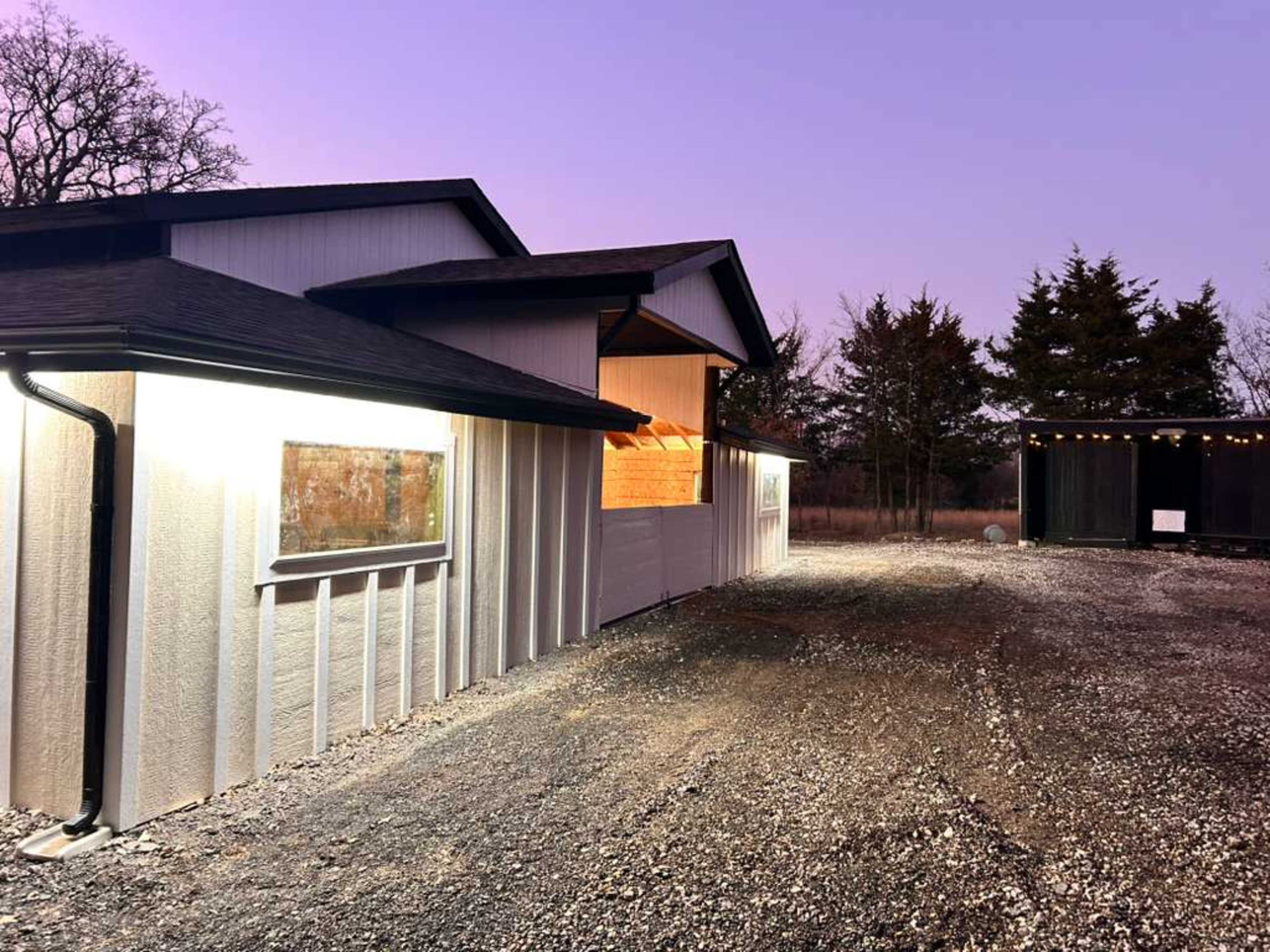 A modern house with a gravel driveway is illuminated by soft lights against a twilight sky, surrounded by trees.