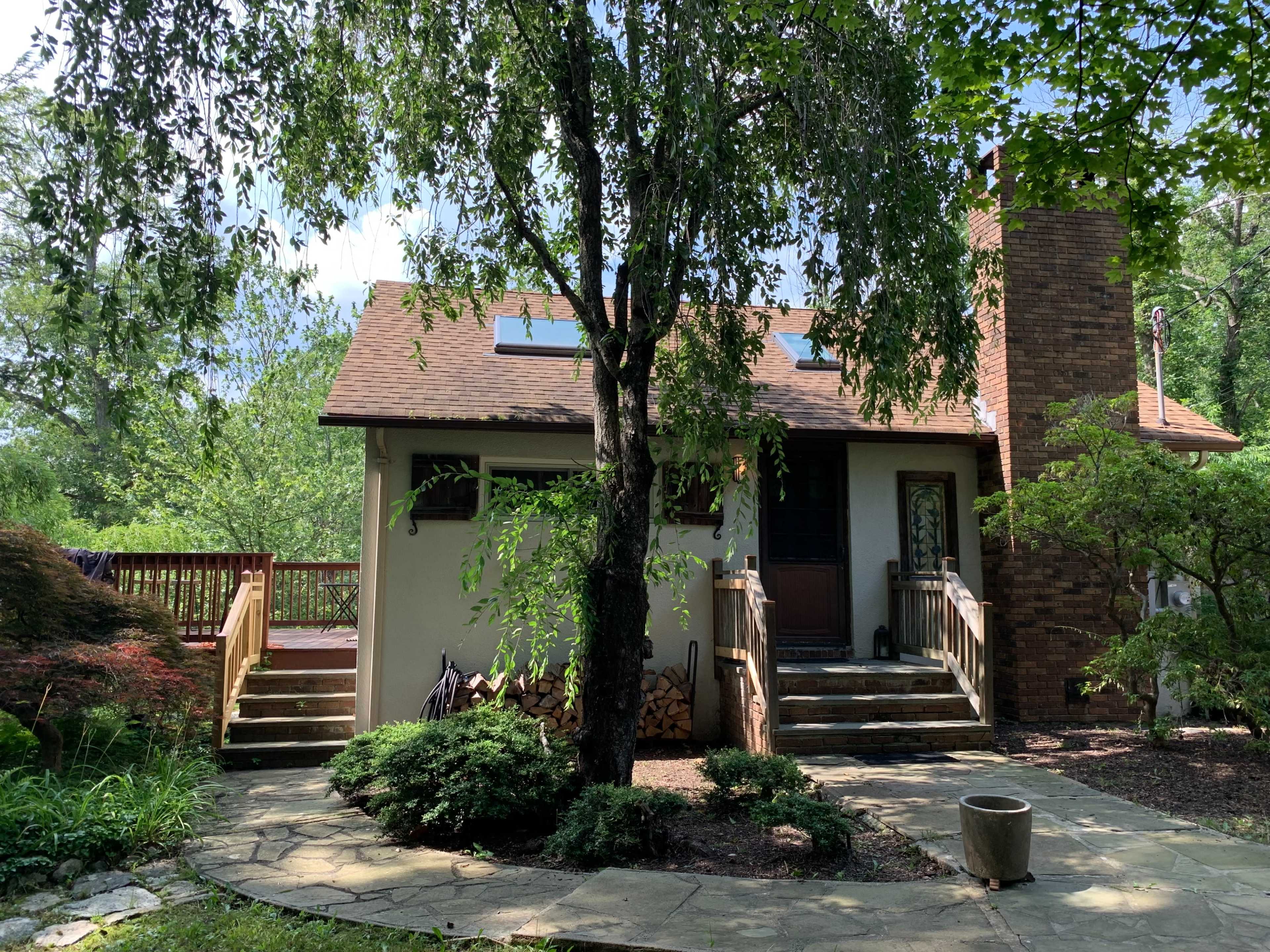A small house with a sloped roof and a brick chimney is surrounded by greenery and features a wooden deck in the front.