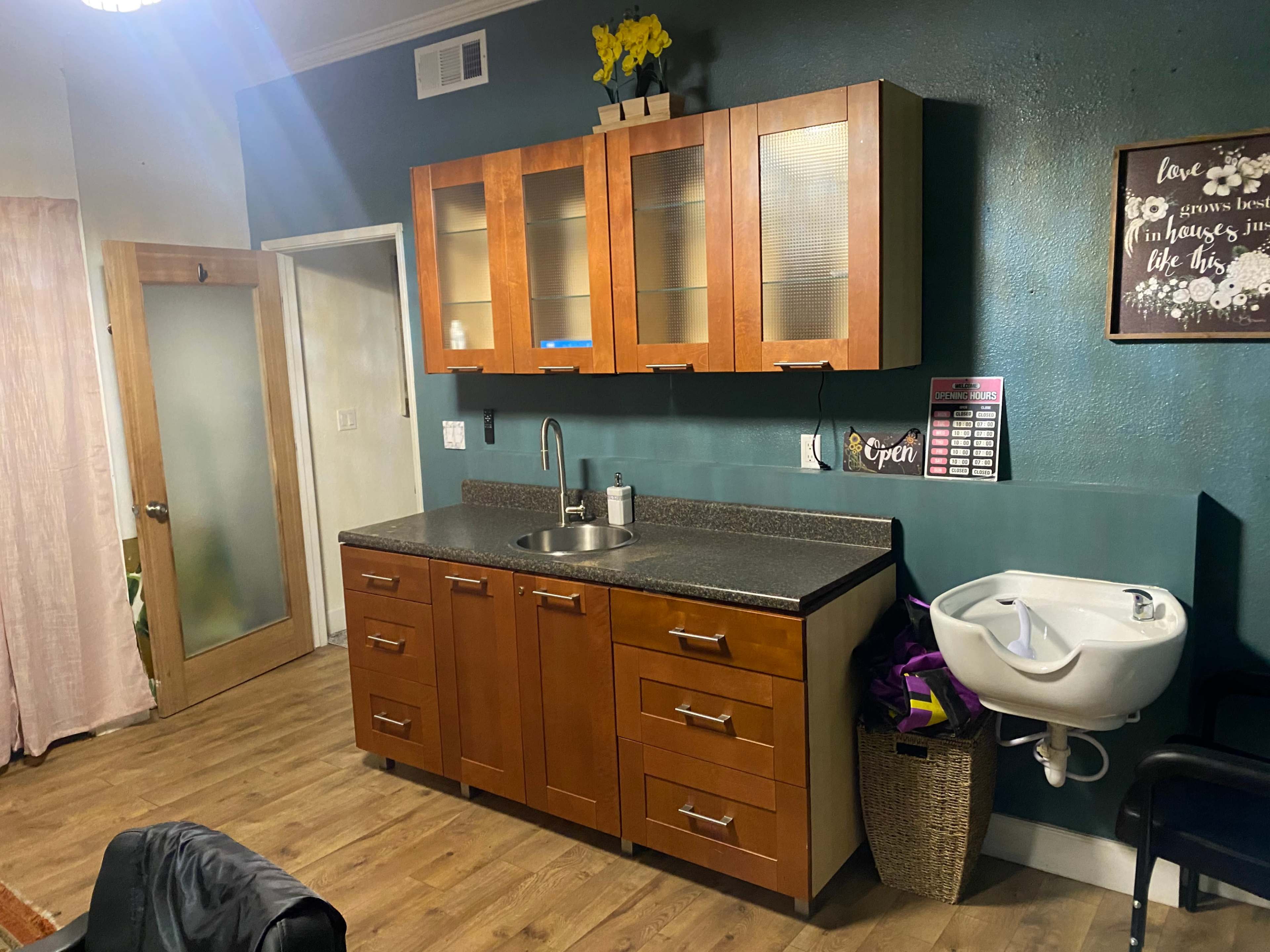 A small room featuring a wooden cabinet with glass doors, a sink, and a wall-mounted washbasin, along with a chair and a decorative sign.