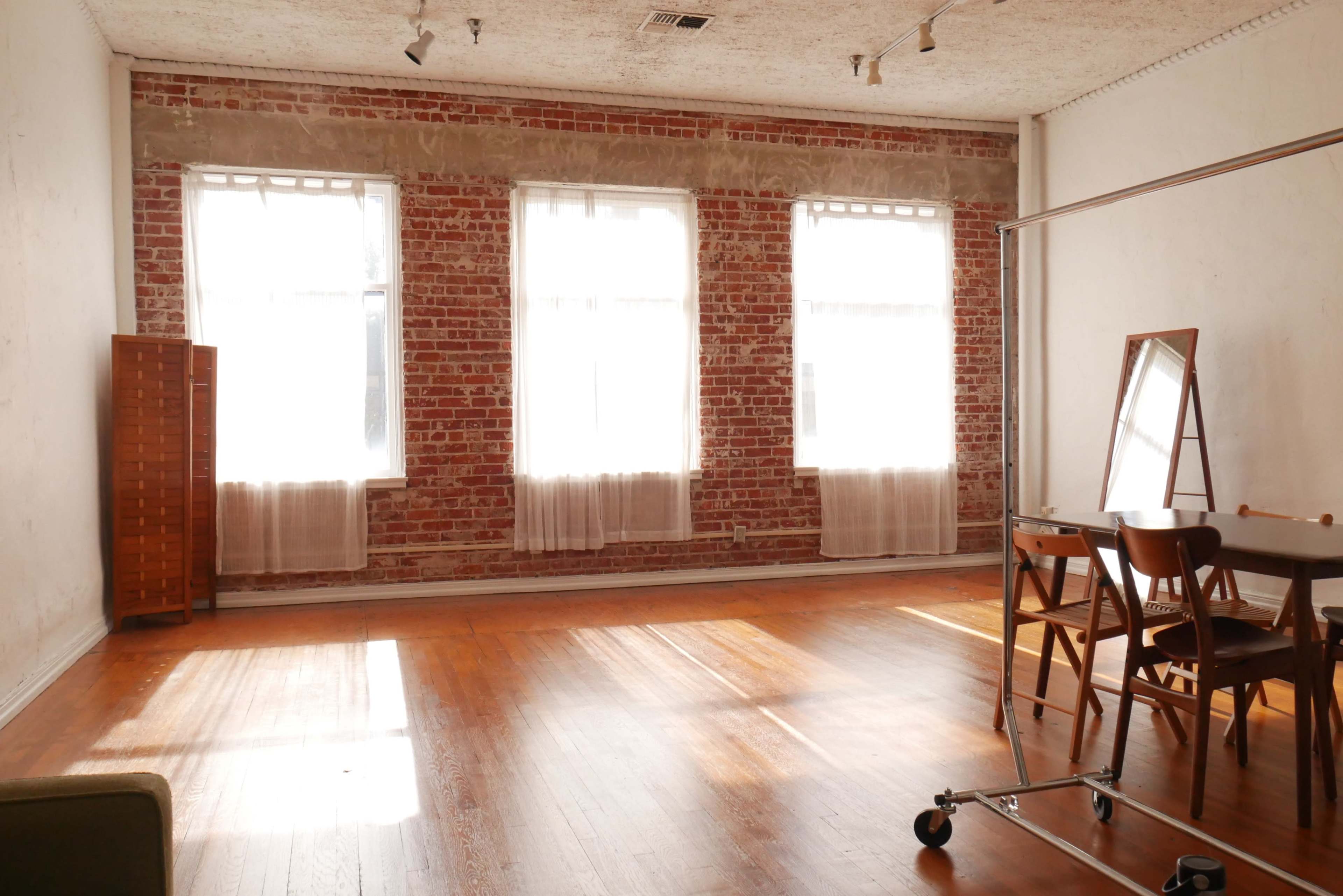 A spacious room with exposed brick walls, large windows, wooden flooring, and a table with chairs.