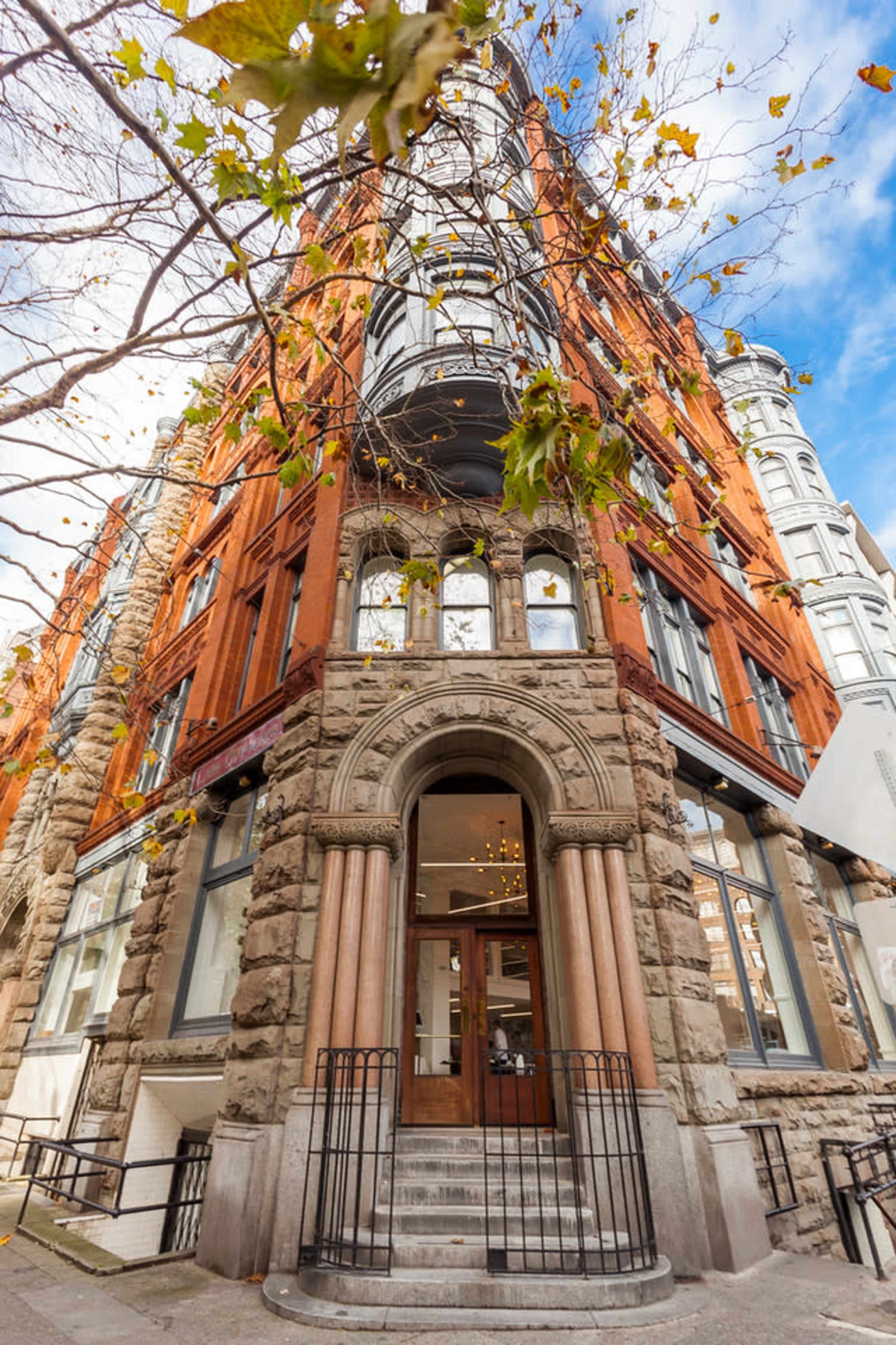 The image shows a historic building with a red brick facade and stone accents at a street corner, featuring large windows and decorative architectural elements.