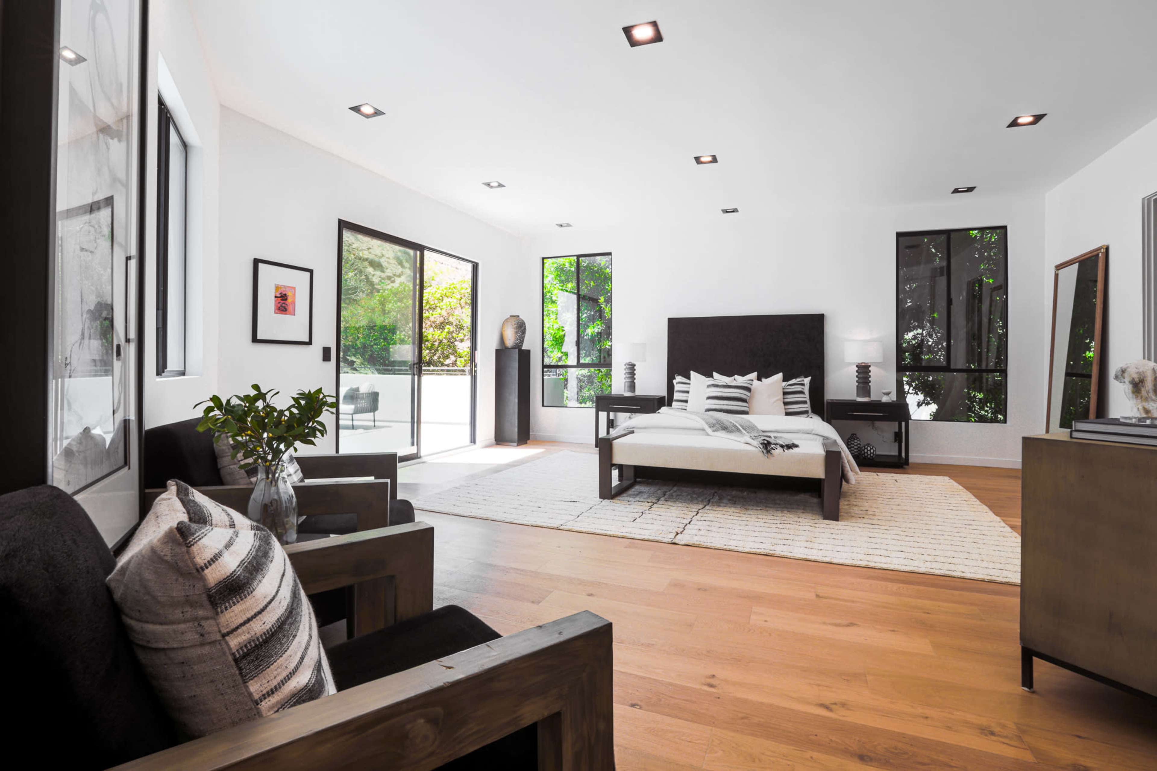 A modern bedroom with a large bed, minimalist decor, wooden flooring, and large windows letting in natural light.