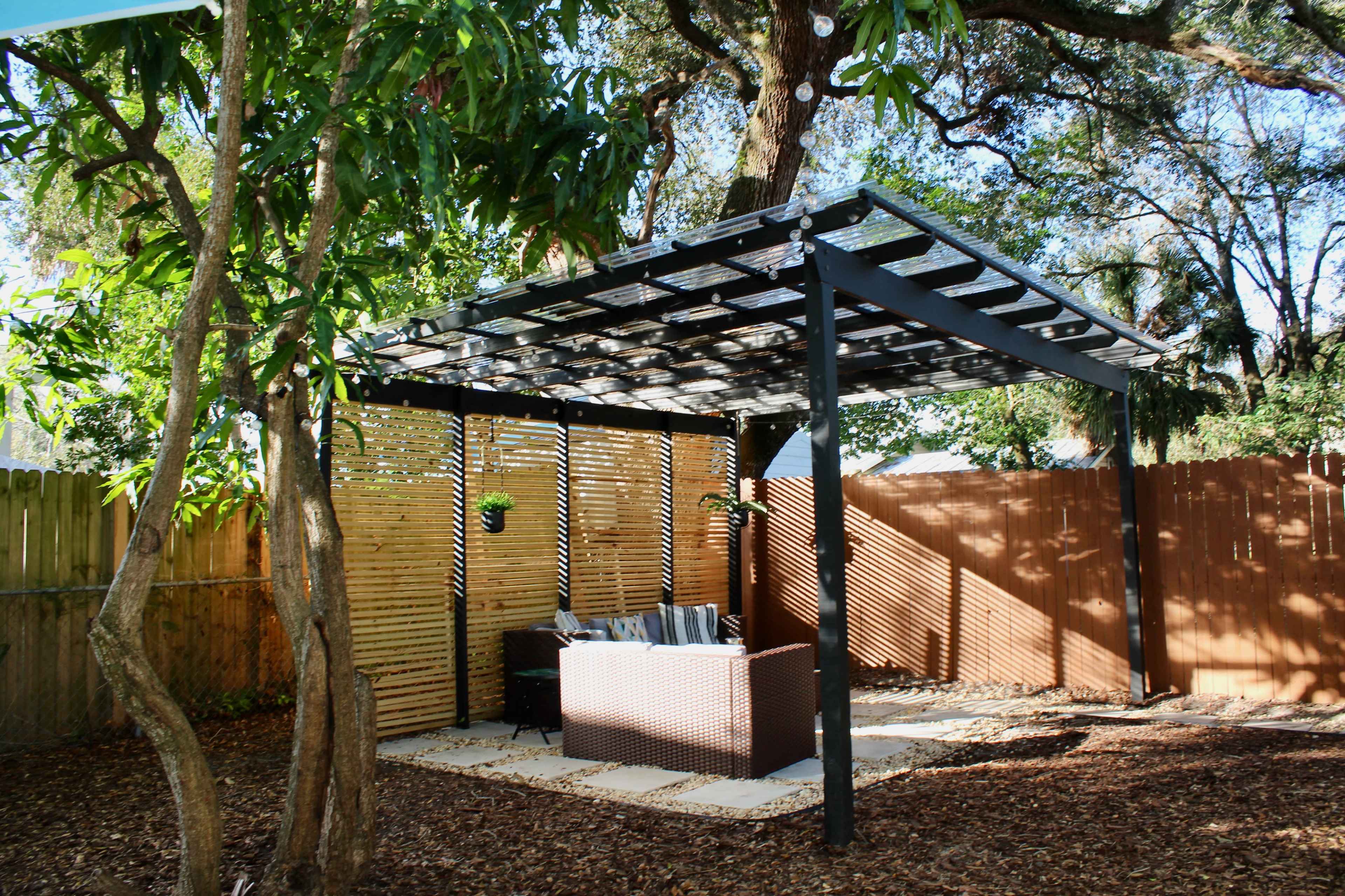 The image shows a shaded outdoor seating area with a pergola, surrounded by trees and a wooden fence.