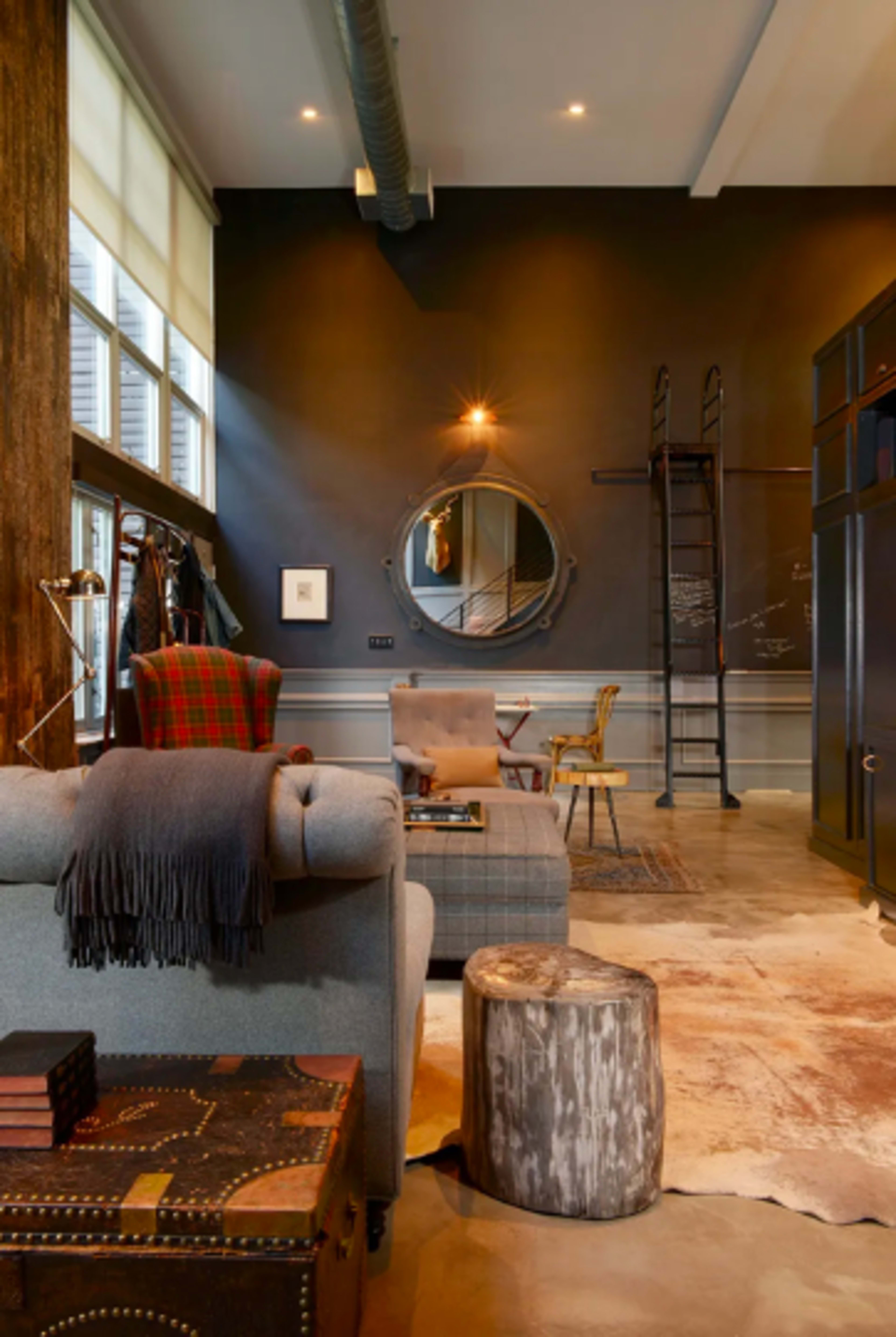The image shows a stylish interior living space with a gray sofa, a round mirror on the dark wall, a wooden stool, and various decorative items scattered on the floor.
