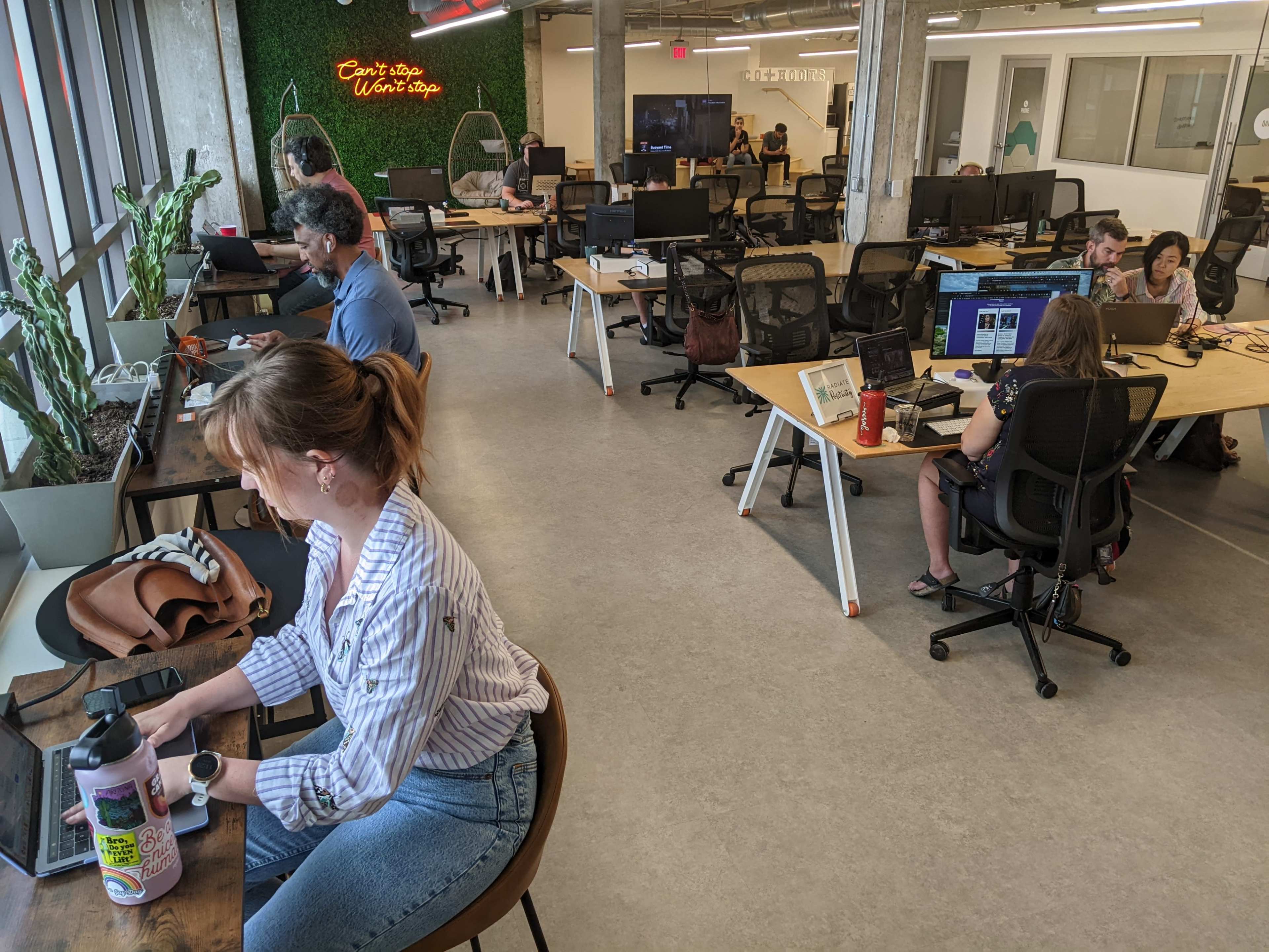 A modern office space features several people working at desks with computers, plants, and collaborative areas.