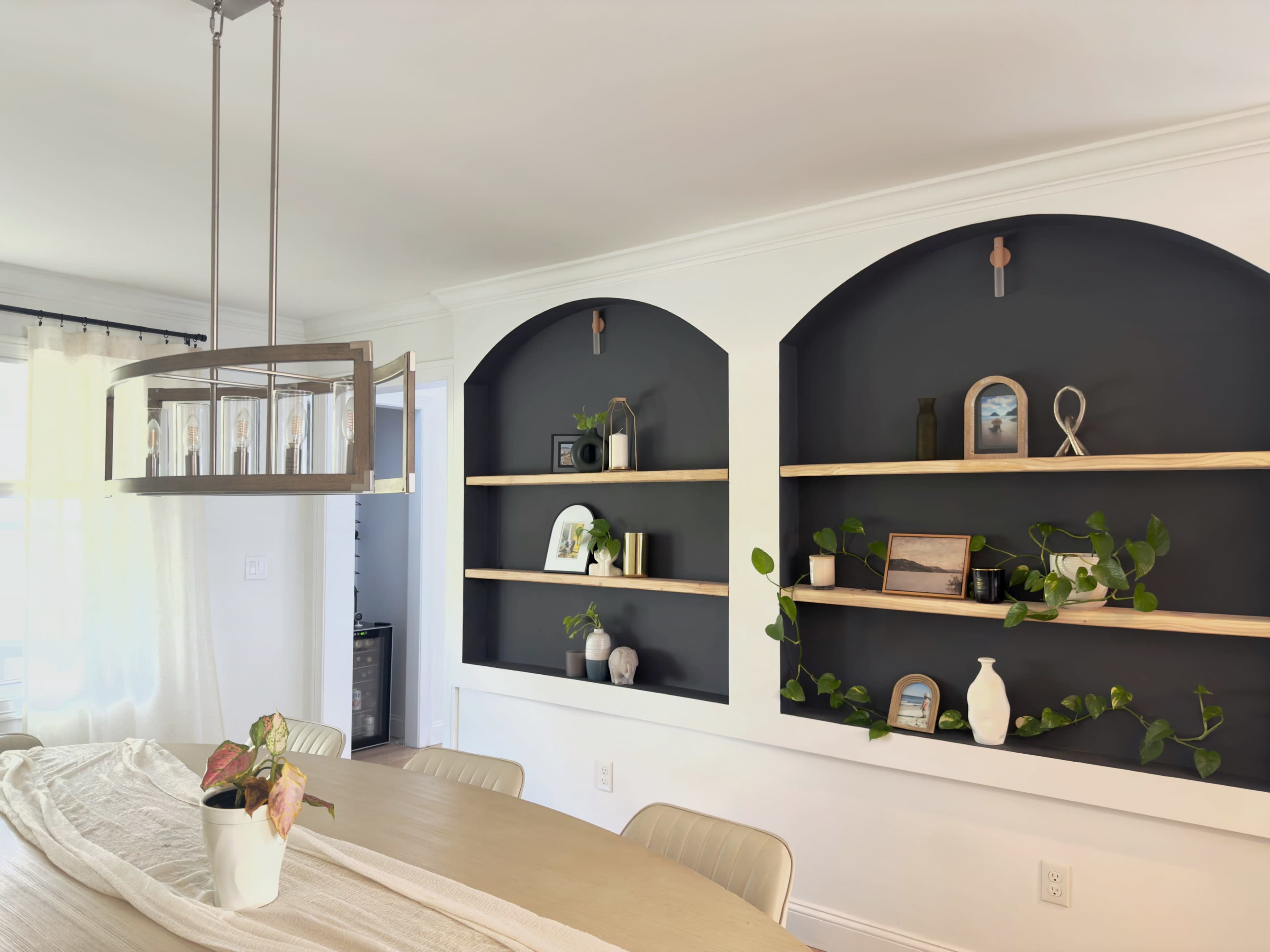 The image shows a modern dining area with a wooden table, a pendant light fixture, and built-in shelves featuring decorative items against a dark wall.