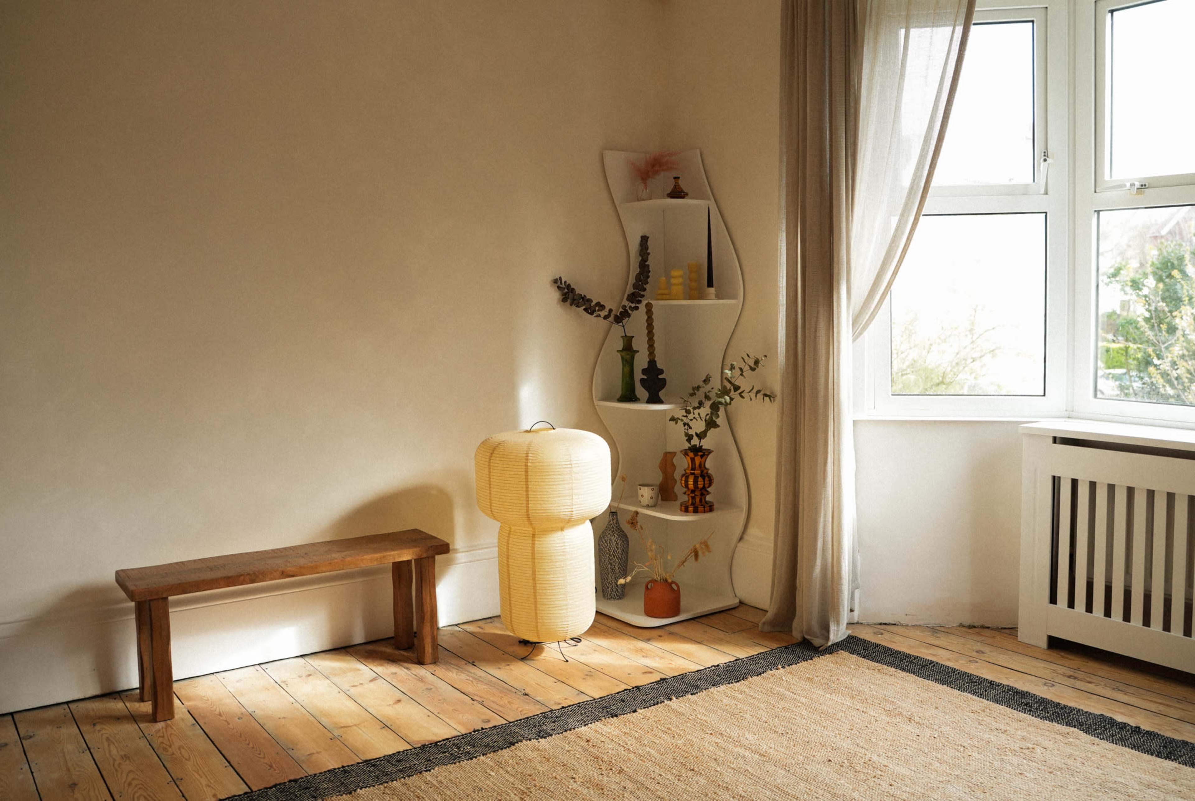 The image depicts a minimalistic room featuring a wooden bench, a lamp, a shelf with decorative items, and a large window letting in natural light.