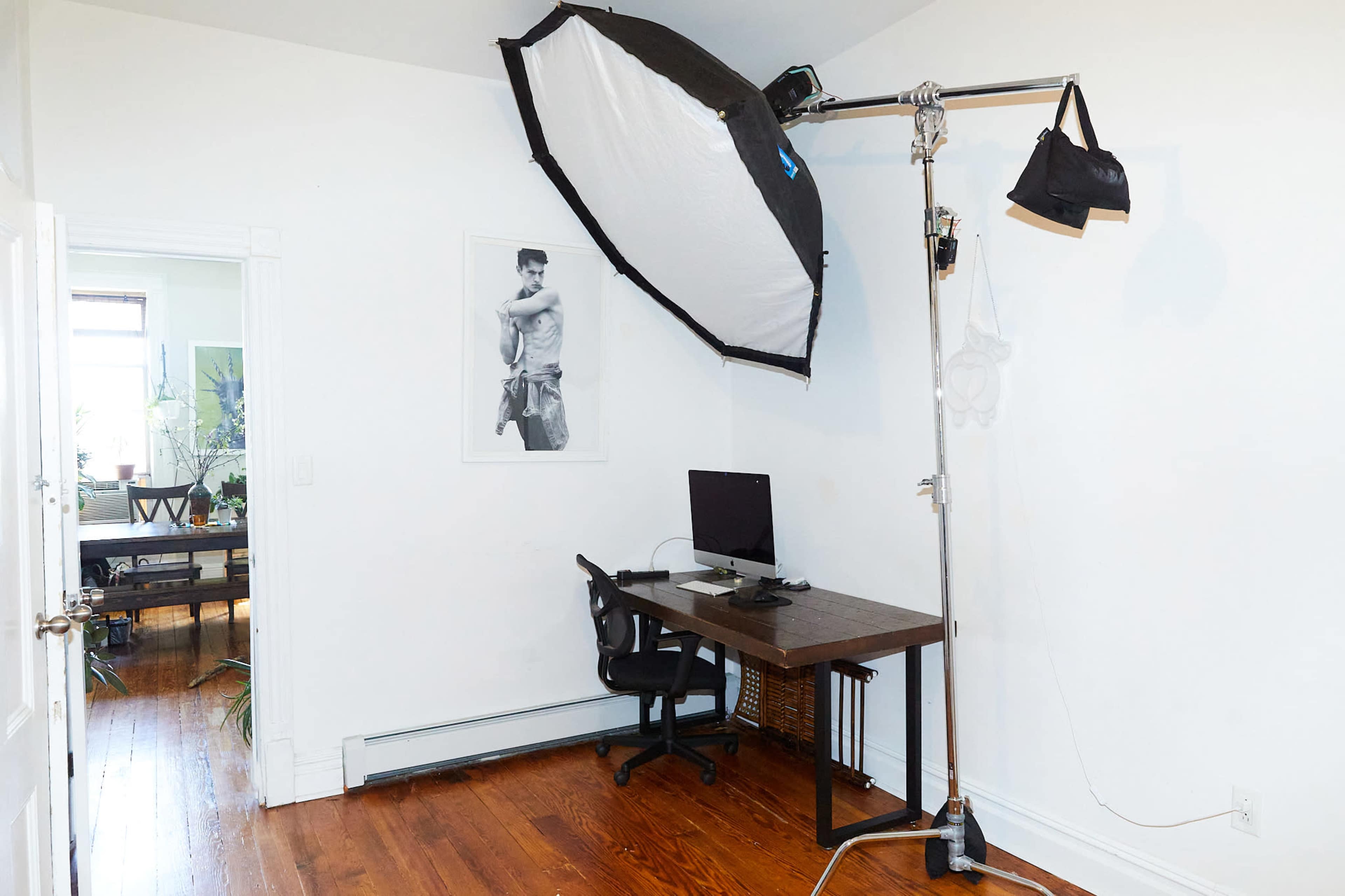 The image shows a home office setup featuring a desk with a computer, a large softbox lighting setup on a stand, and a wall with a framed poster.