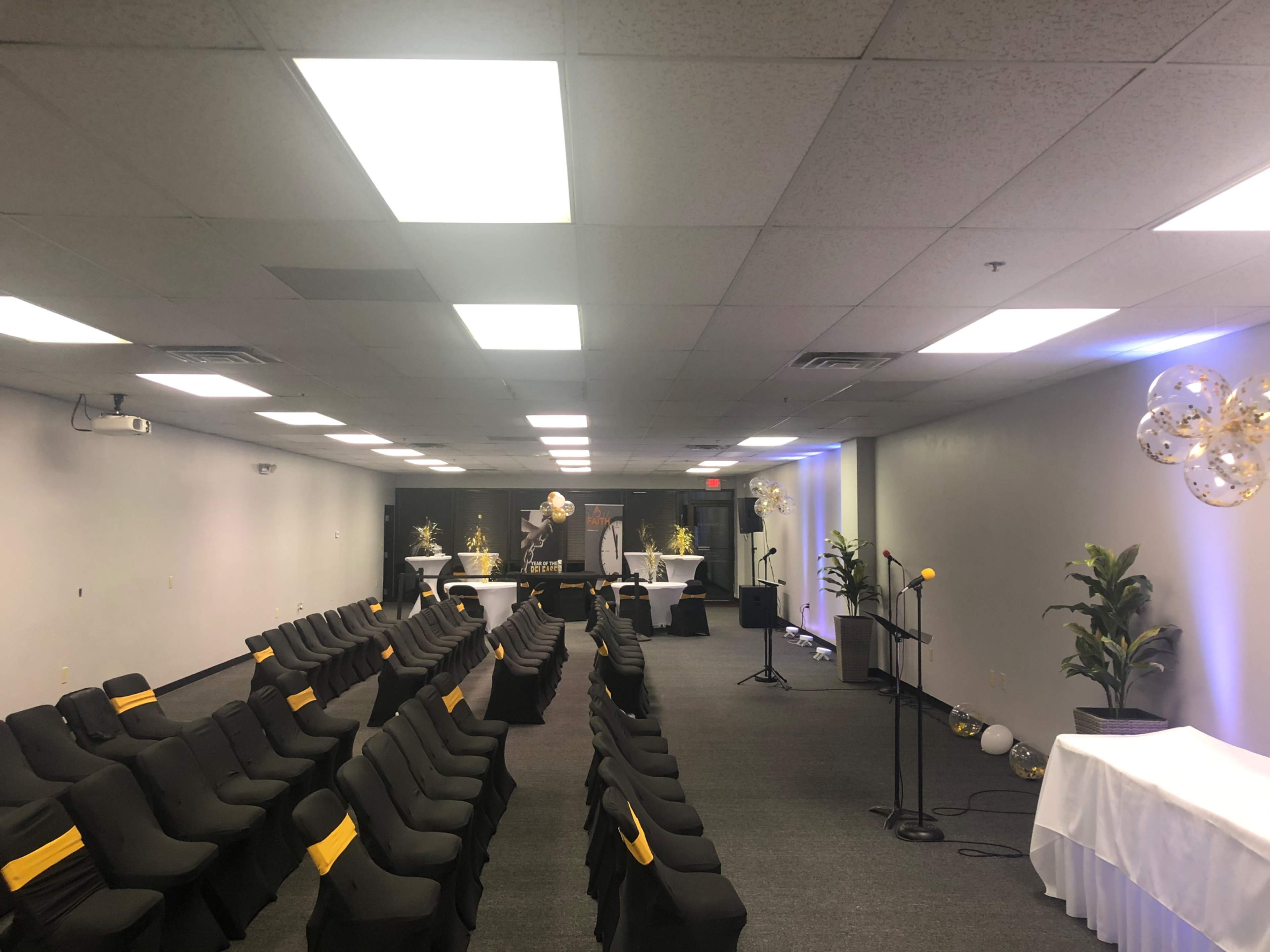 The image shows a modern event space set up with rows of black chairs featuring yellow accents, a stage with a microphone, and decorative elements including plants and tables.