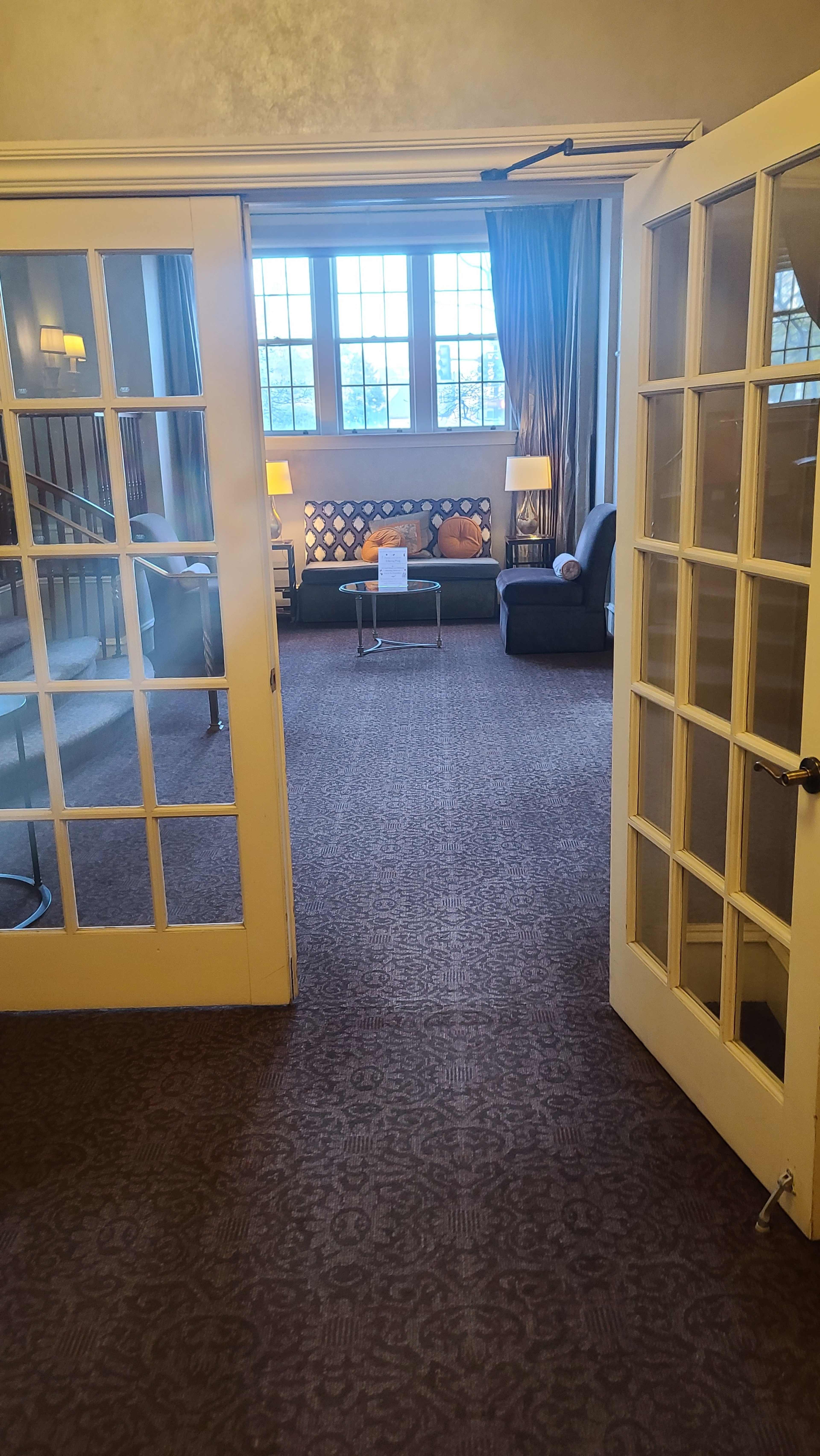 The image shows an interior space with two glass panel doors leading into a room that features a seating area with a couch and chairs, bathed in natural light from large windows.