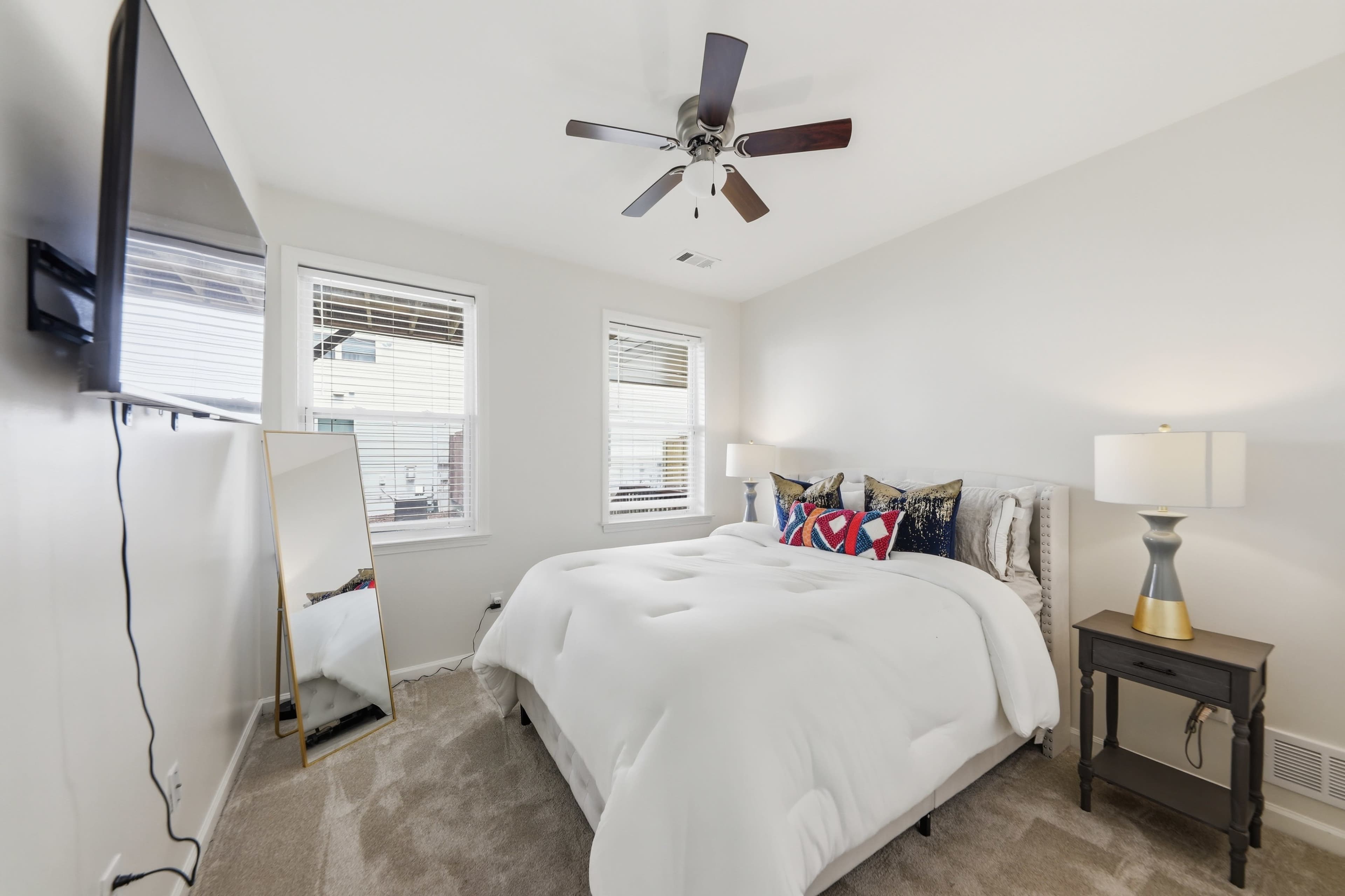 A well-lit bedroom features a queen-sized bed with white bedding, a large mirror, and a TV mounted on the wall.