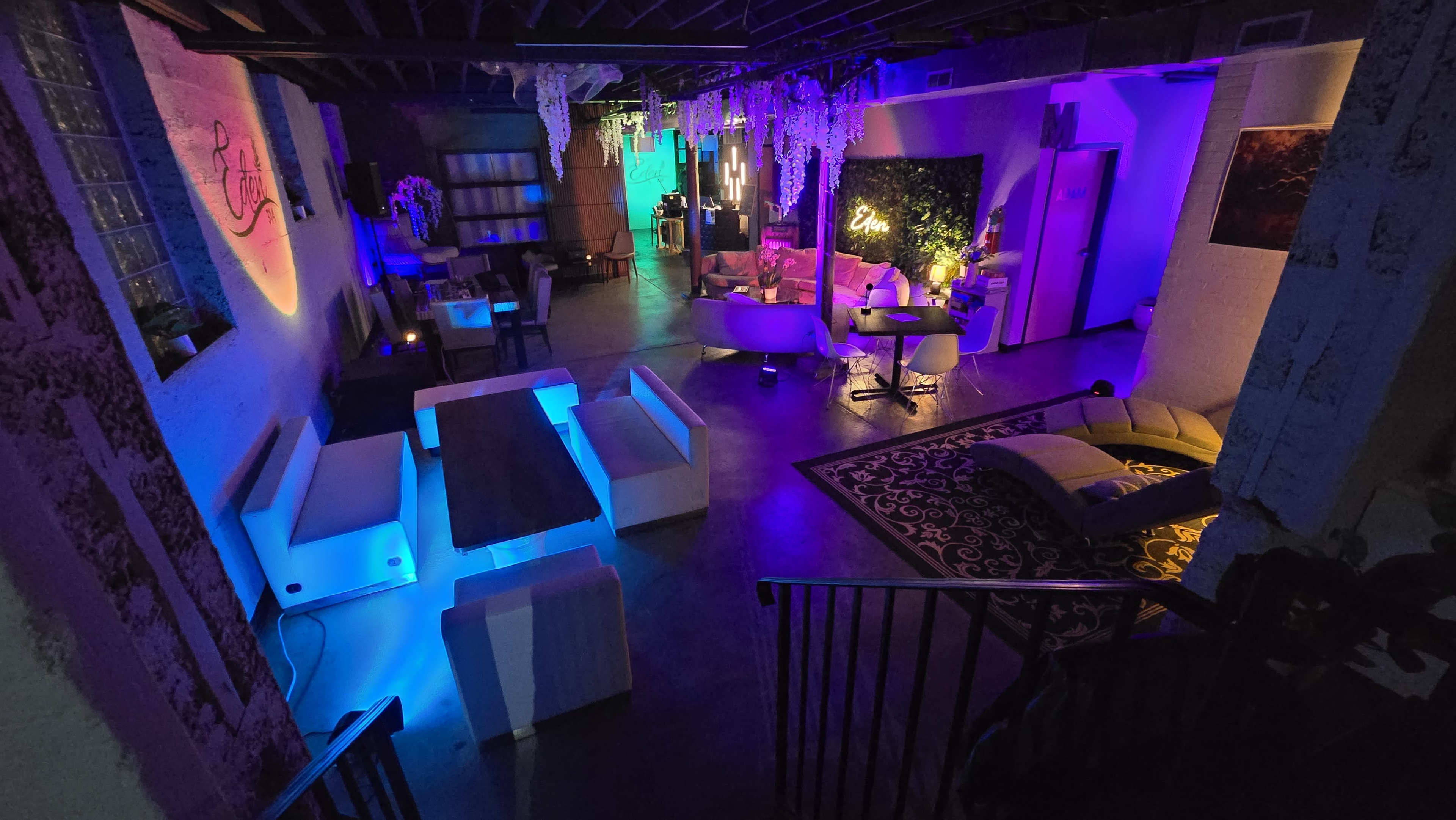 The image shows a modern lounge area with stylish furniture, illuminated by colorful lights and decorated with plants.