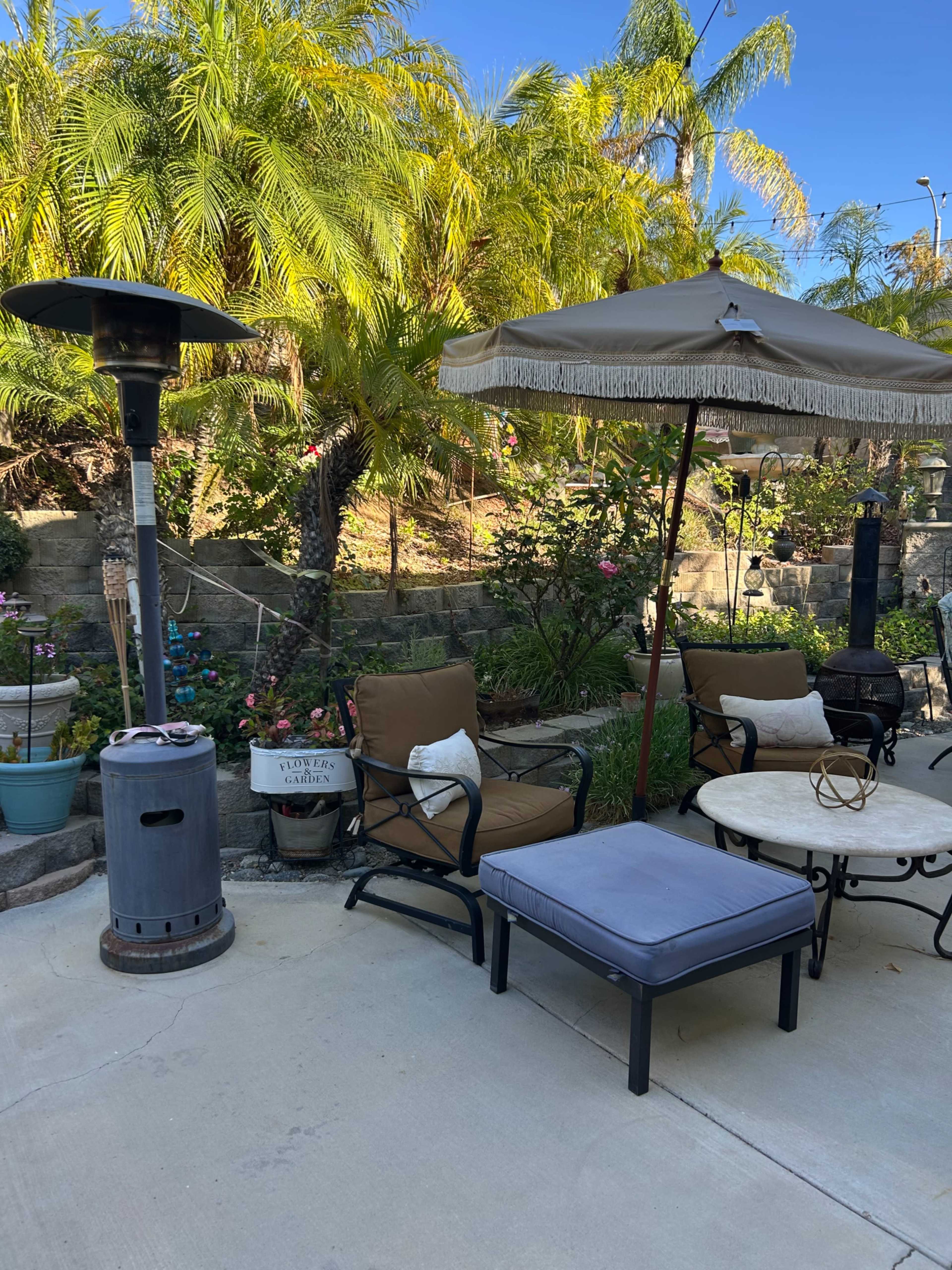 The image shows a backyard patio area with two chairs, a table, a patio umbrella, a heater, and various plants in the background.