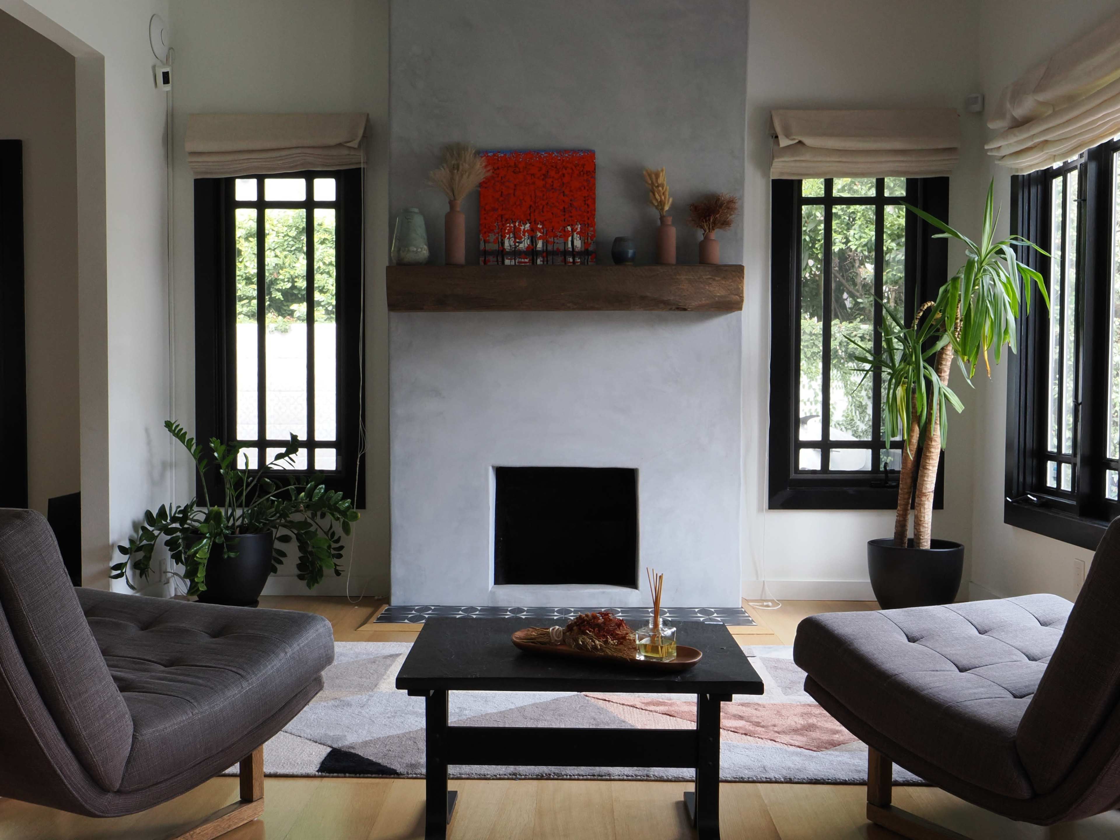 A modern living room features two gray armchairs, a dark coffee table, a minimalist fireplace with a wooden mantel, and large windows allowing natural light to enter.