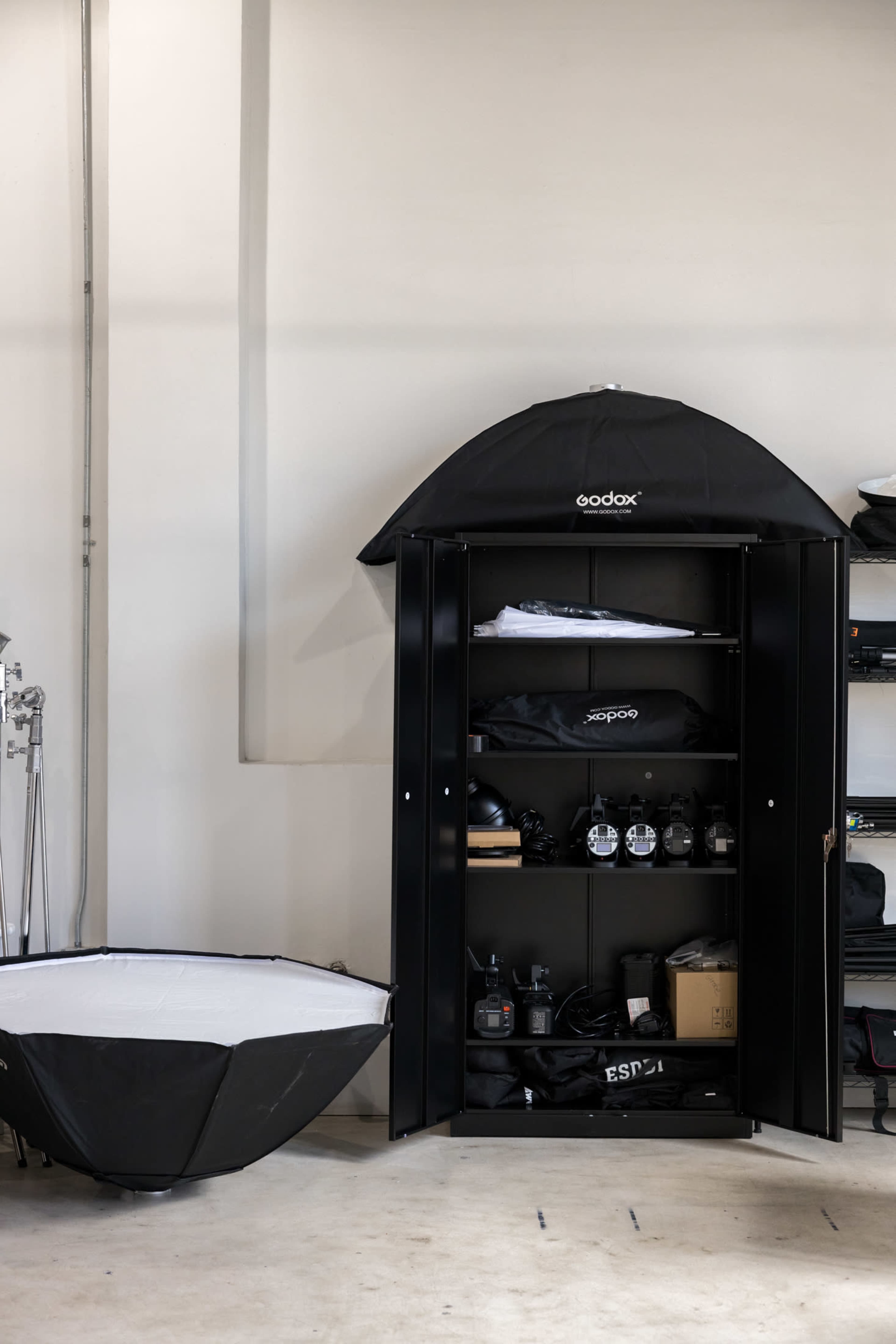 A black storage cabinet is open, displaying various photography equipment, while a collapsible light modifier sits on the floor next to it.