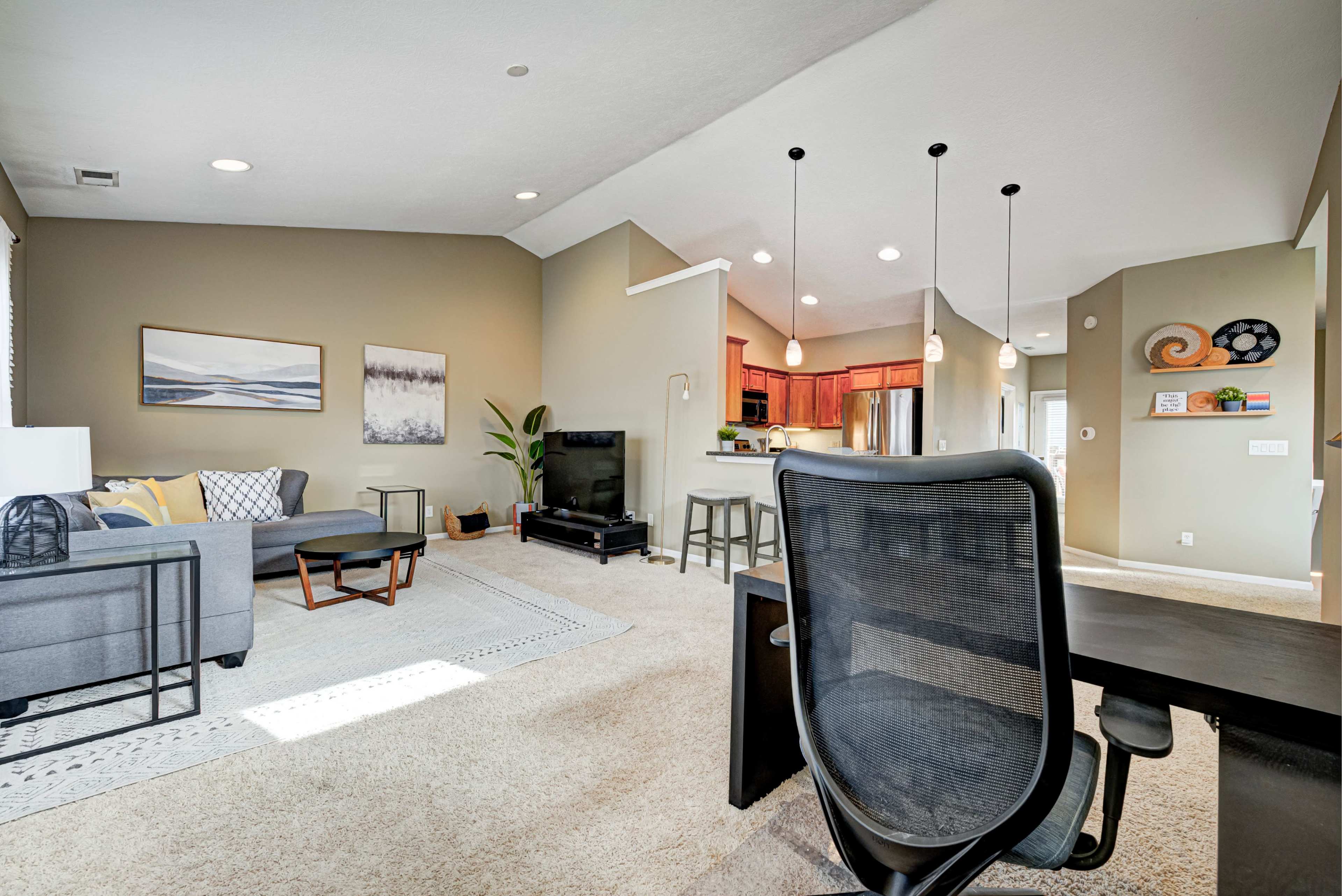 A modern living room features a gray sofa, a black TV stand, and a desk, with an open kitchen visible in the background.