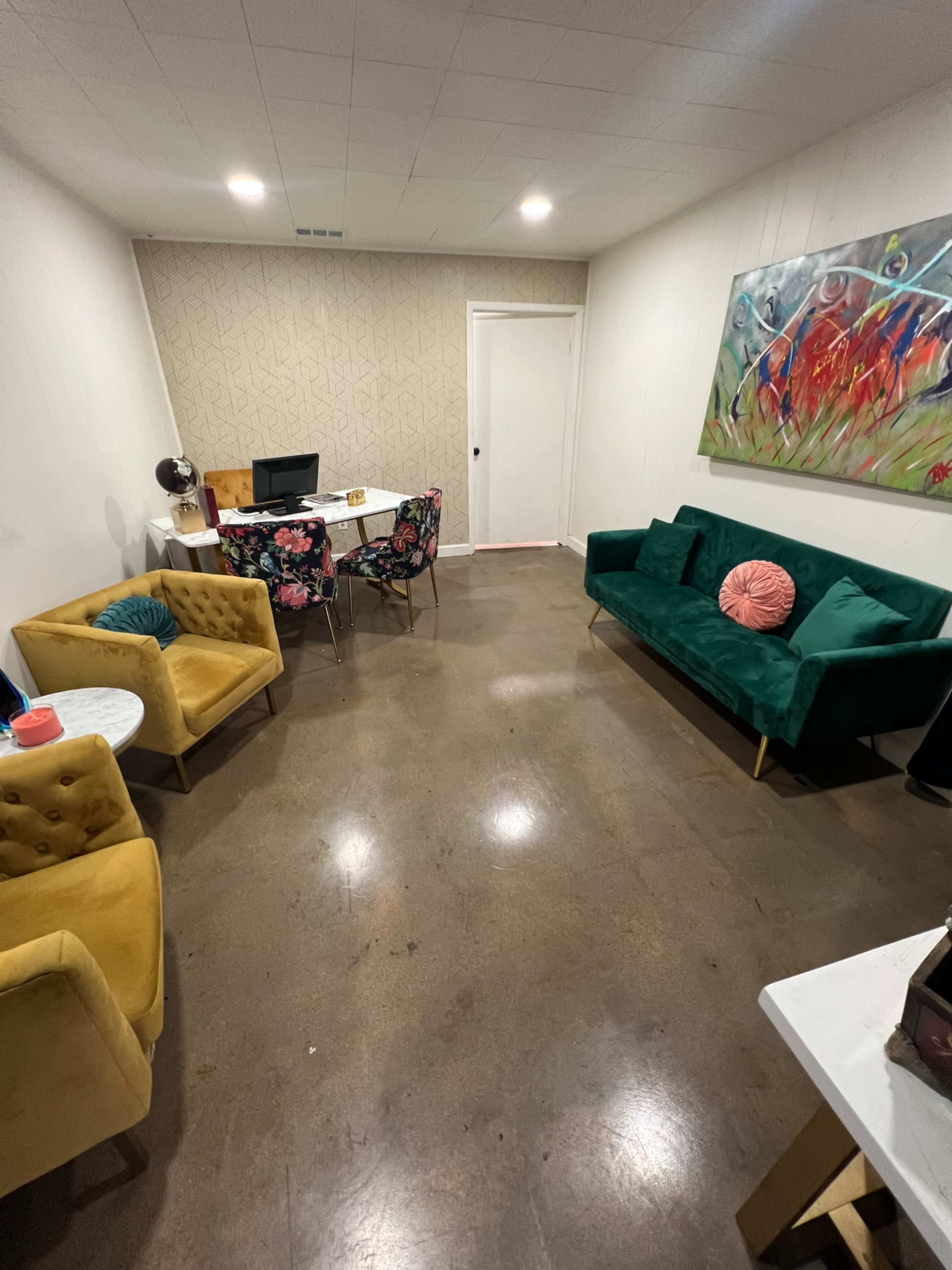 The image shows a small office space featuring a desk and computer, surrounded by a green sofa, yellow armchairs, and a floral-patterned table.