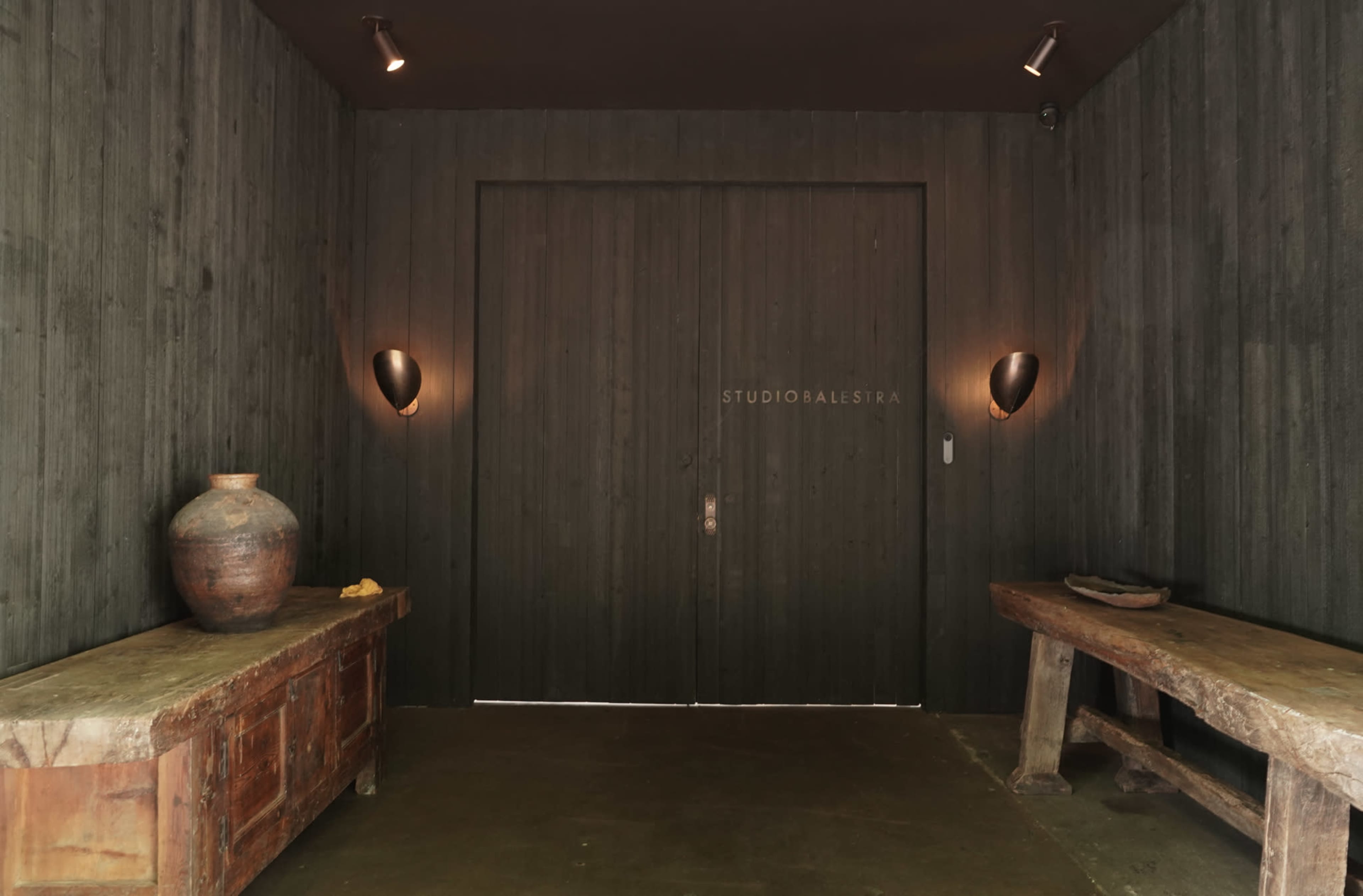 The image depicts a dimly lit hallway featuring wooden walls, two wall-mounted lights, and rustic furniture including two benches and a large pottery jar.