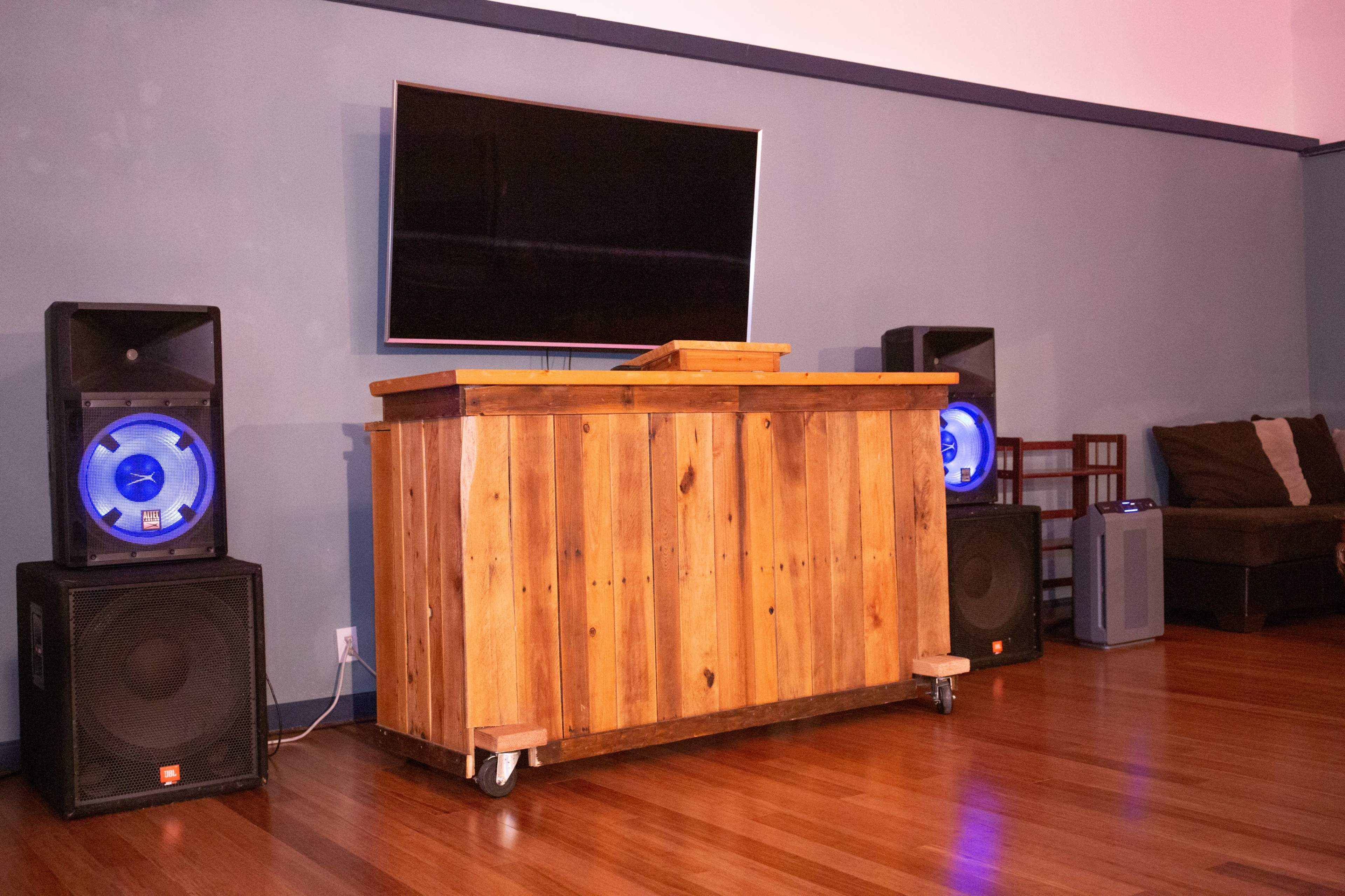 A wooden bar with a flat-screen television mounted above it, flanked by two large speakers on either side, in a room with wood flooring and a couch in the background.