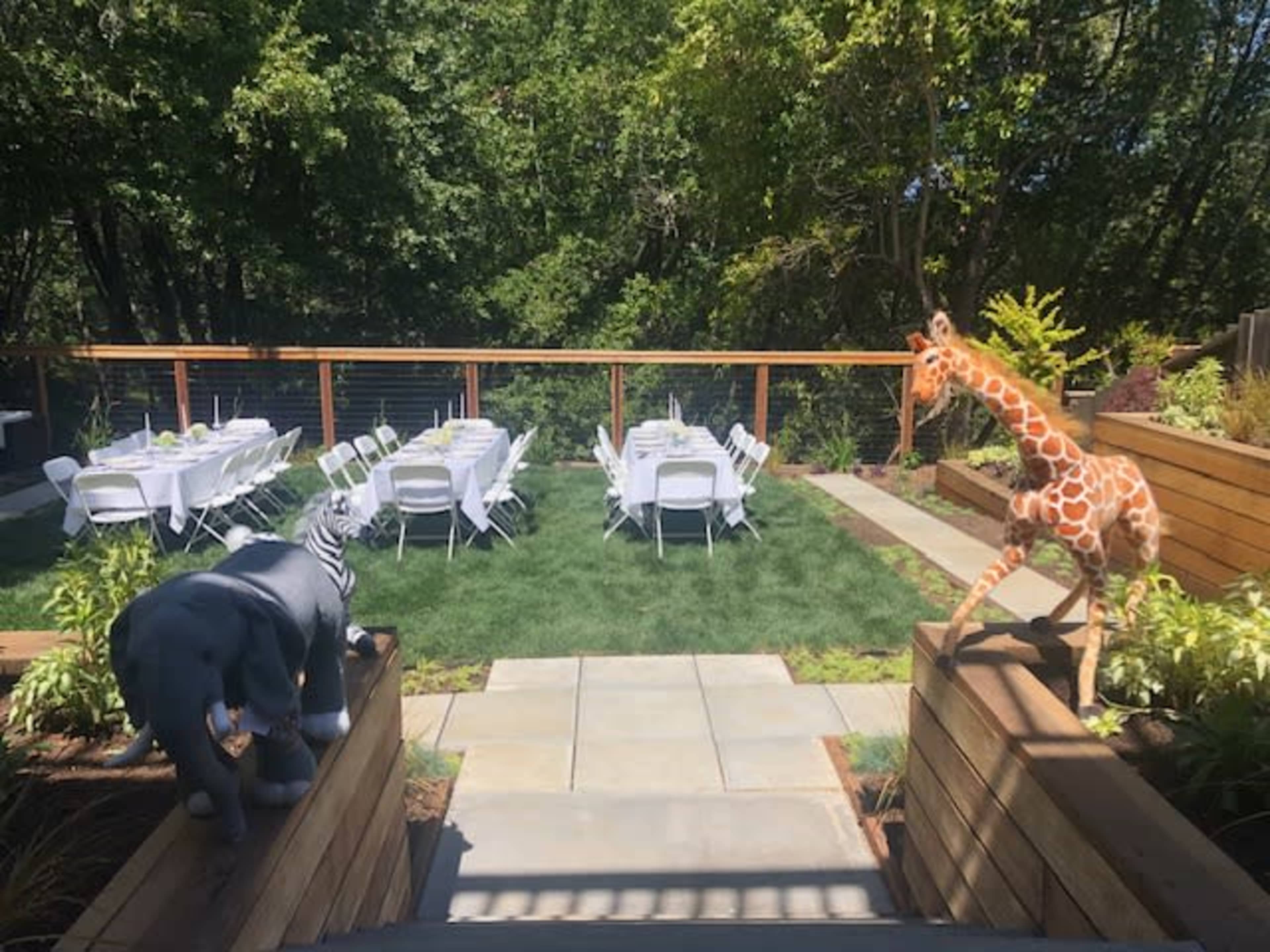 A backyard event space featuring rows of white tables and chairs arranged on green grass, framed by decorative animal statues of a giraffe and an elephant.