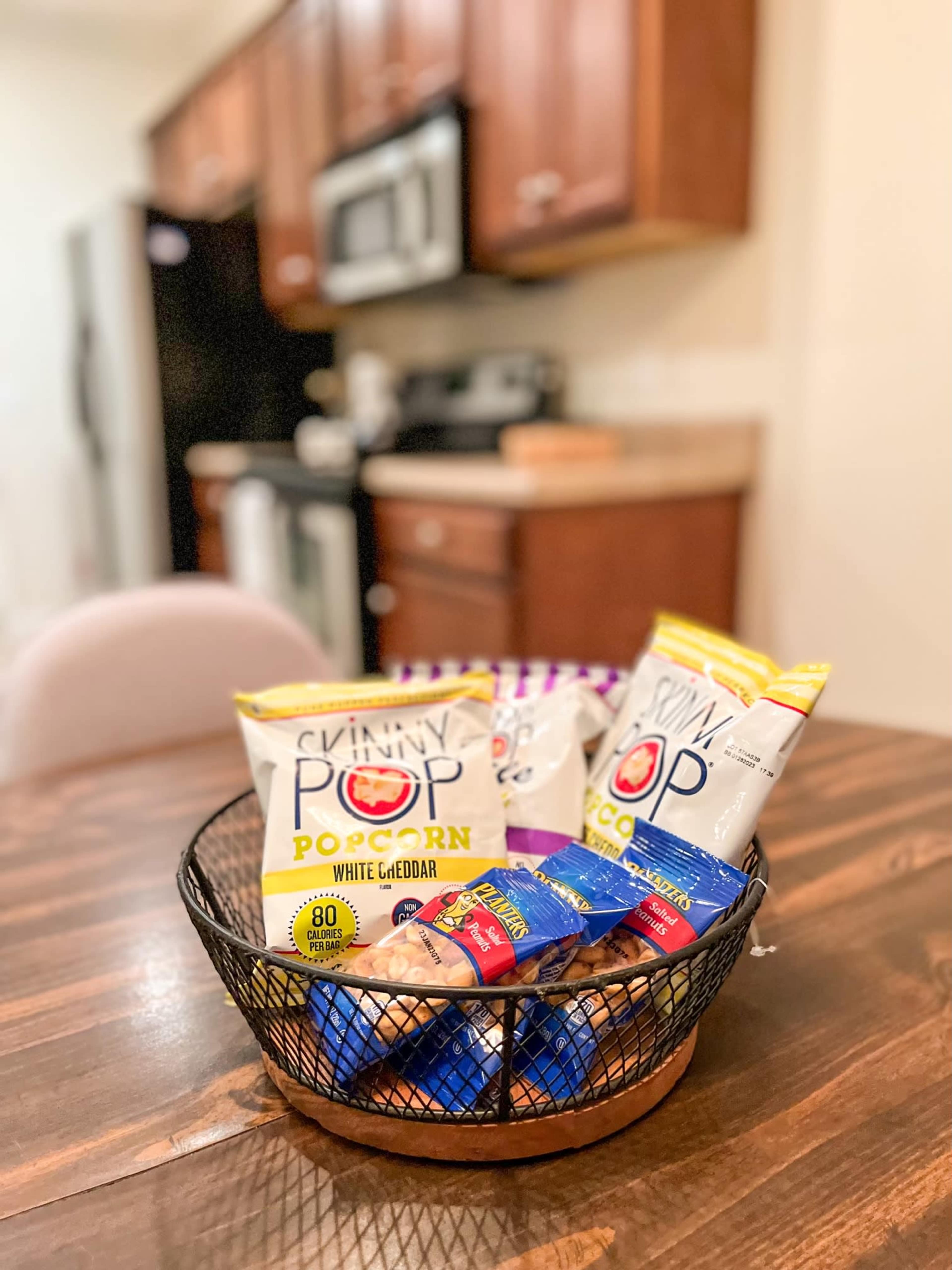 A wire basket filled with packs of SkinnyPop popcorn and snacks sits on a wooden table in a kitchen.