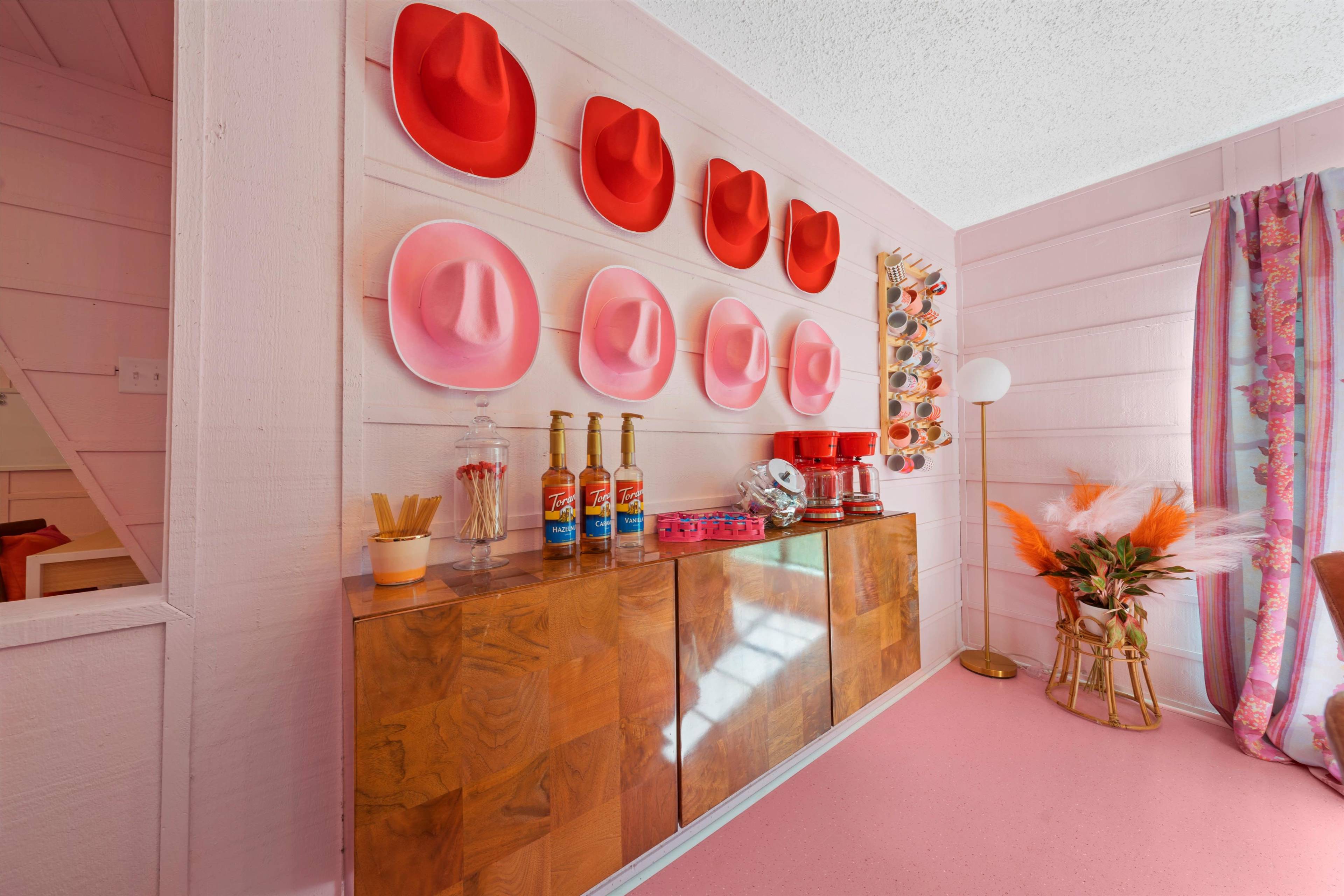 A brightly colored interior with a pink floor, a wooden cabinet displaying bottles and snacks, and a wall adorned with red cowboy hats.